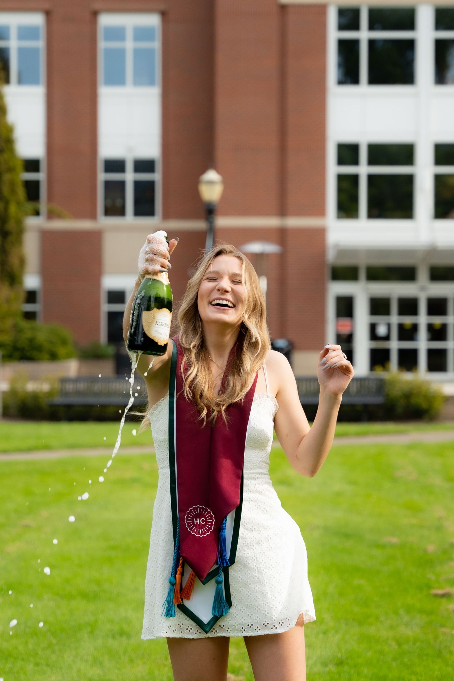 Graduate in white dress and burgundy stole celebrates with champagne on campus lawn in front of brick building.
