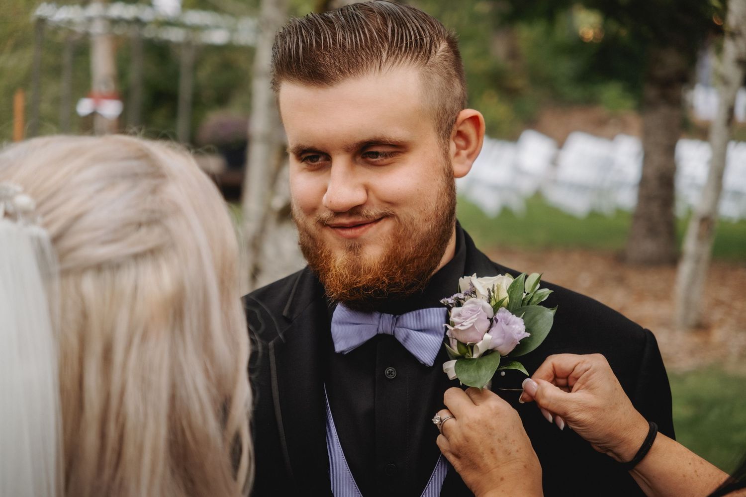 Groomsmen in formal black suits laugh and smile while adjusting boutonnieres during an outdoor wedding.