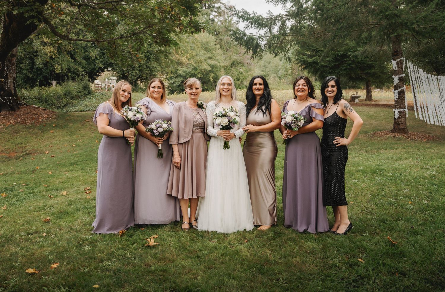 Wedding party group photo with bridesmaids in coordinating dusty purple and mauve dresses on a lawn.