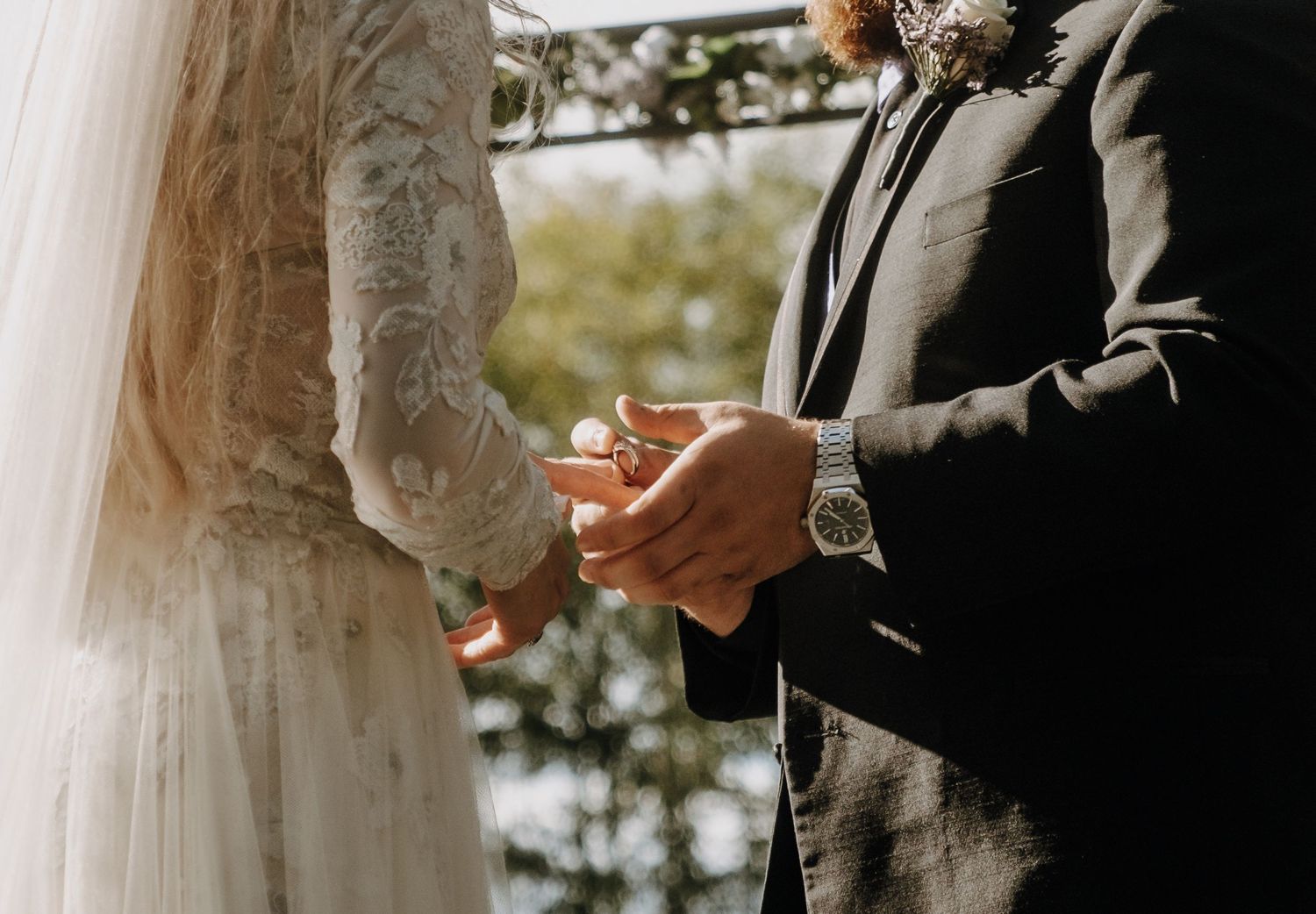 Close-up view of hands exchanging wedding rings during an outdoor ceremony with lace dress and suit details.