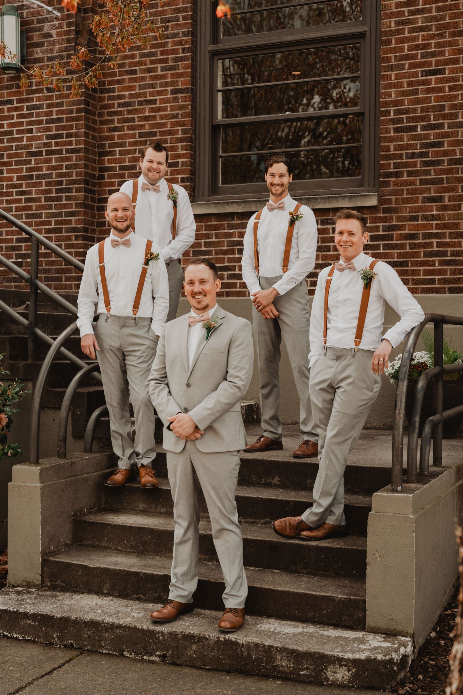 A wedding party in light gray suits and brown leather suspenders poses on brick building steps.