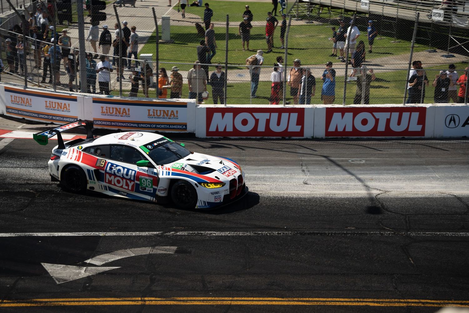 White BMW race car with sponsor decals competes on a racing circuit with Motul advertising banners and spectators visible.