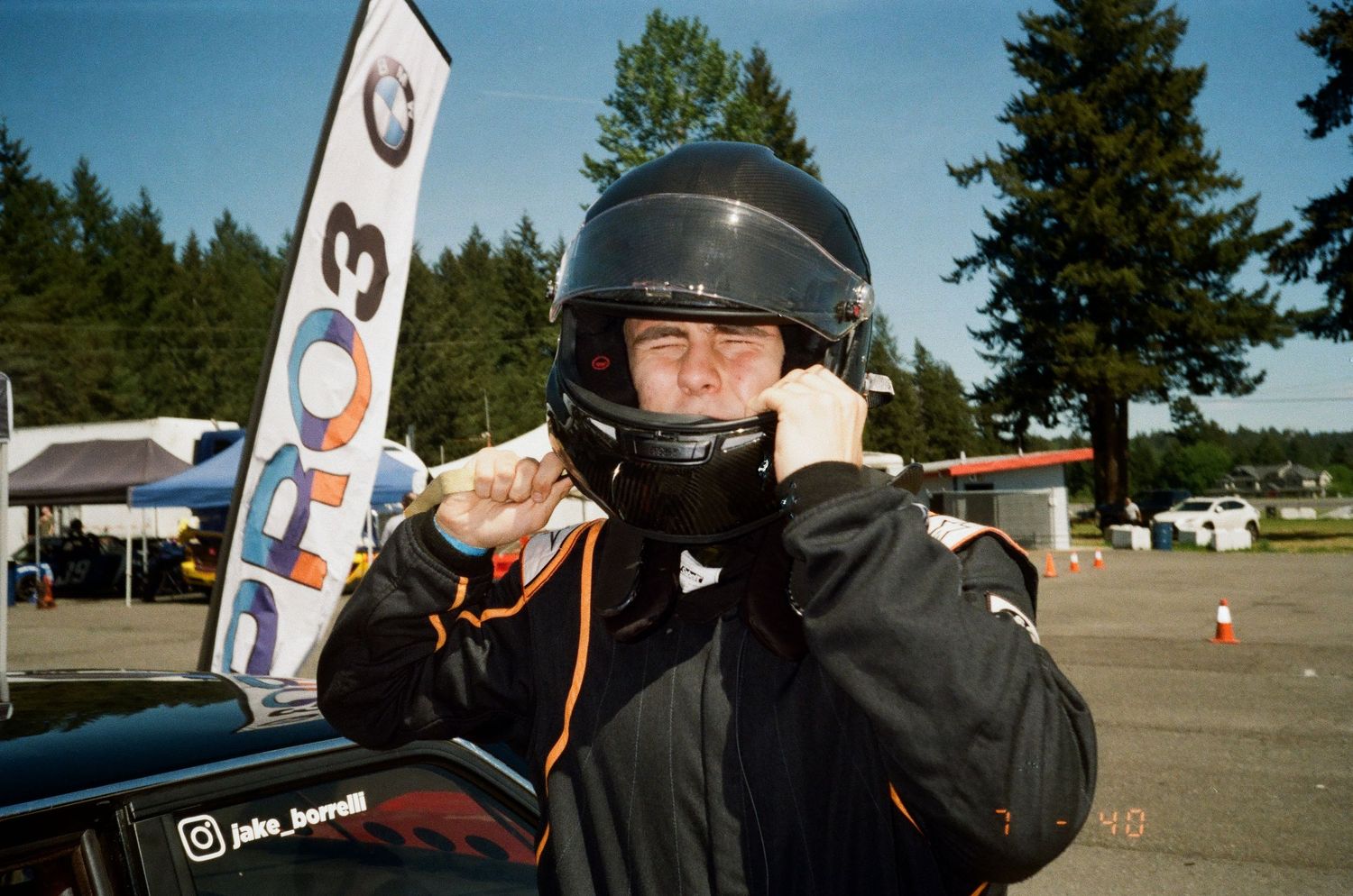 Motorcyclist in black protective gear and helmet near racing event sign adjusts their helmet at outdoor event.