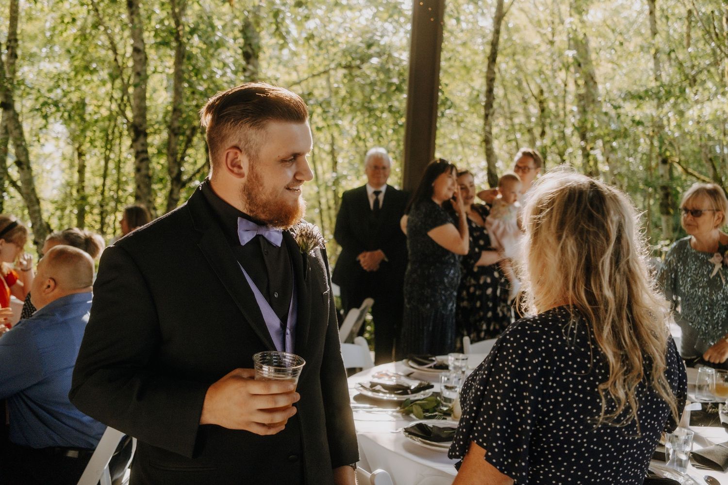 Wedding guests mingle and socialize in a sun-dappled outdoor forest setting.
