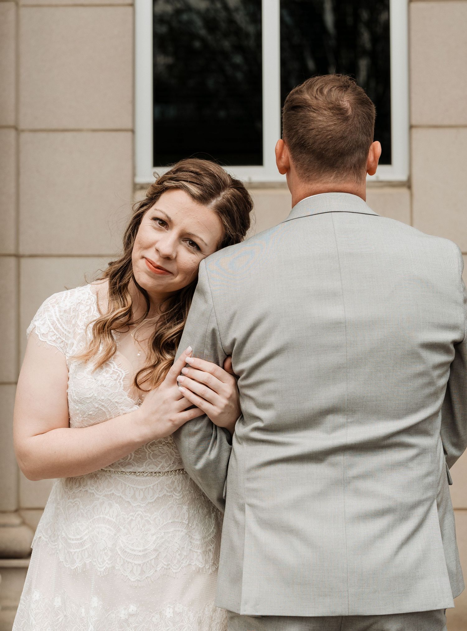 A bride in a lace wedding dress holds onto her partner wearing a light gray suit as they pose by a building.