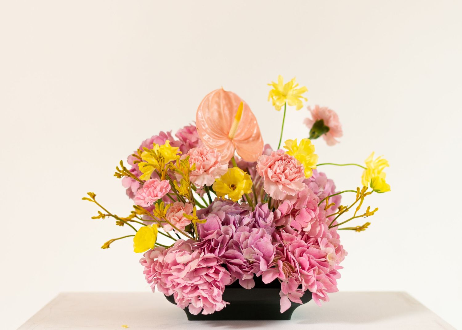 A delicate floral arrangement with pink carnations and yellow daisies in a black container against a neutral backdrop.