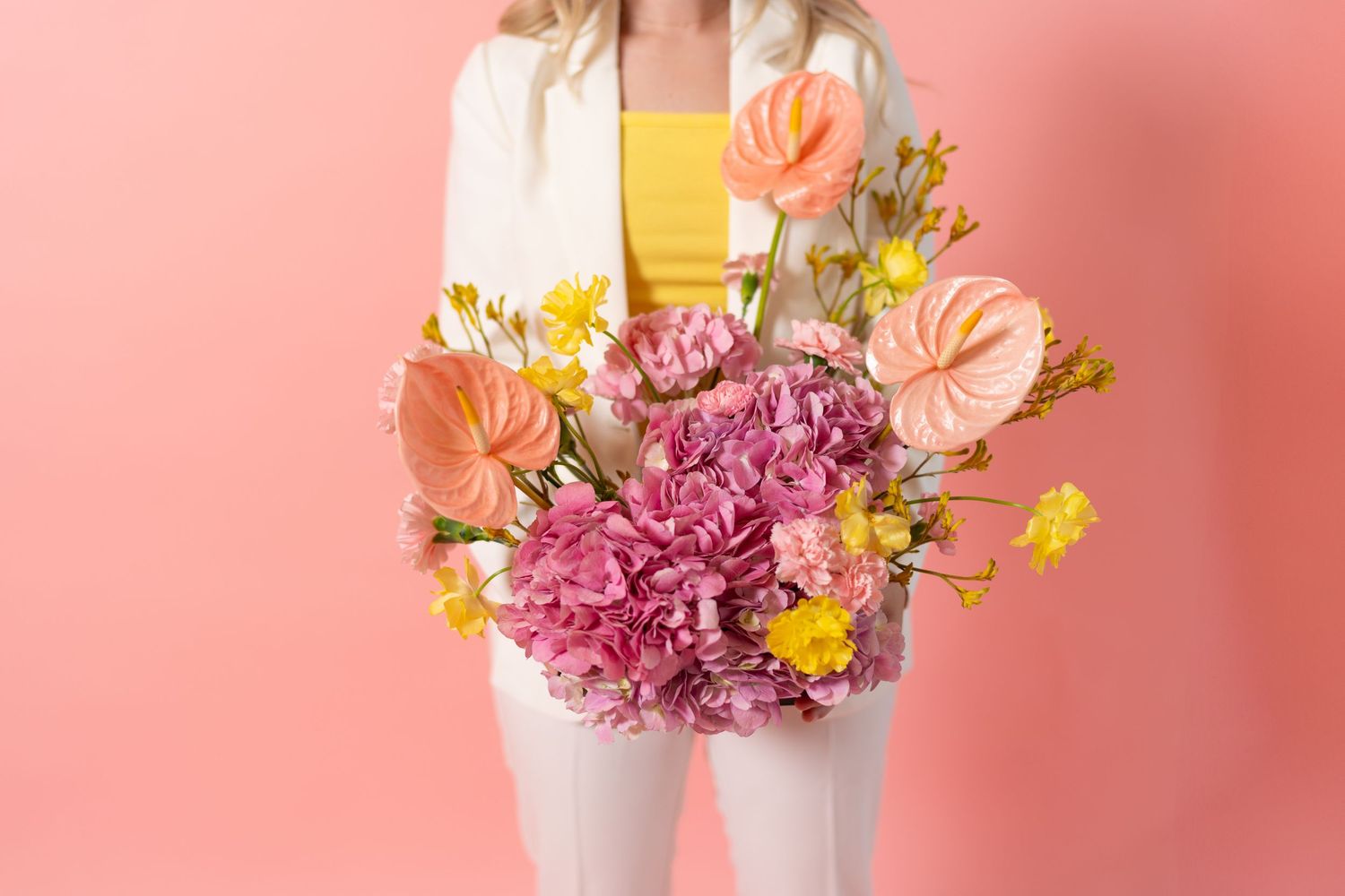 Close-up of colorful floral arrangement with pink hydrangeas, orange anthuriums, and yellow flowers against coral background.