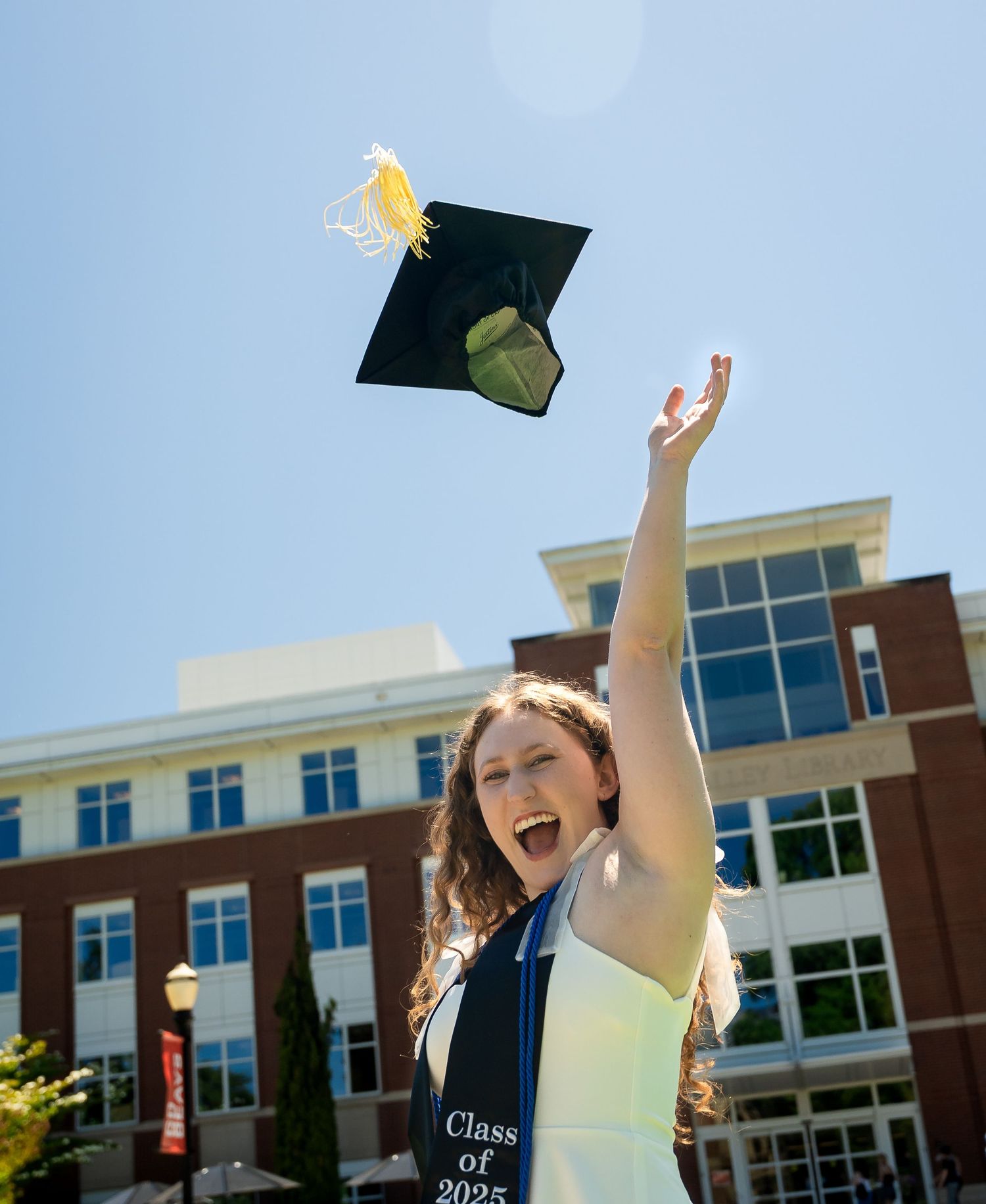 Graduate celebrating and tossing graduation cap in the air outside a brick university building on a sunny day.