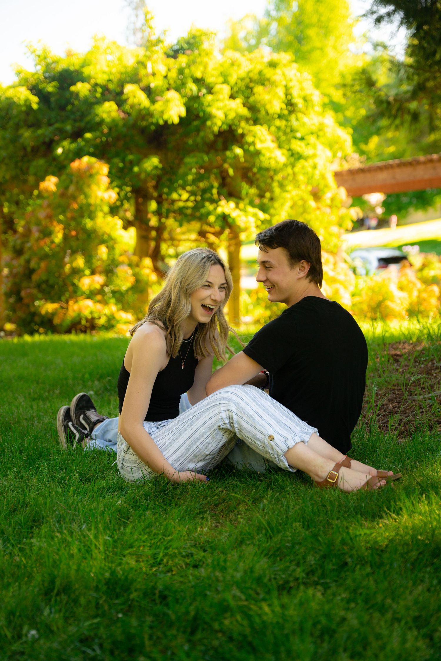 A couple shares intimate laughter while laying on green grass with golden sunlight in the background.