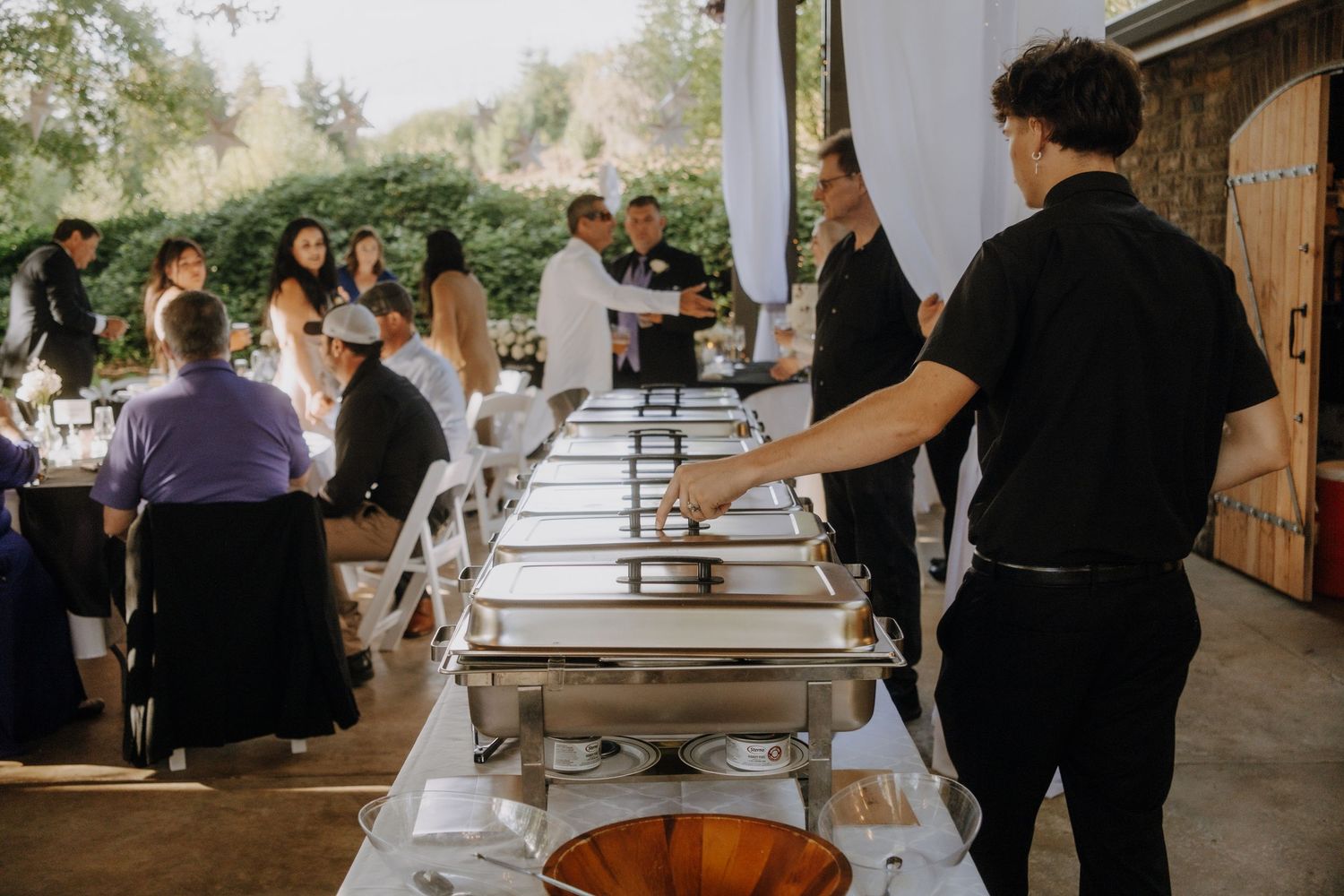 Catering staff sets up buffet service stations at an outdoor event with chafing dishes along white tables.