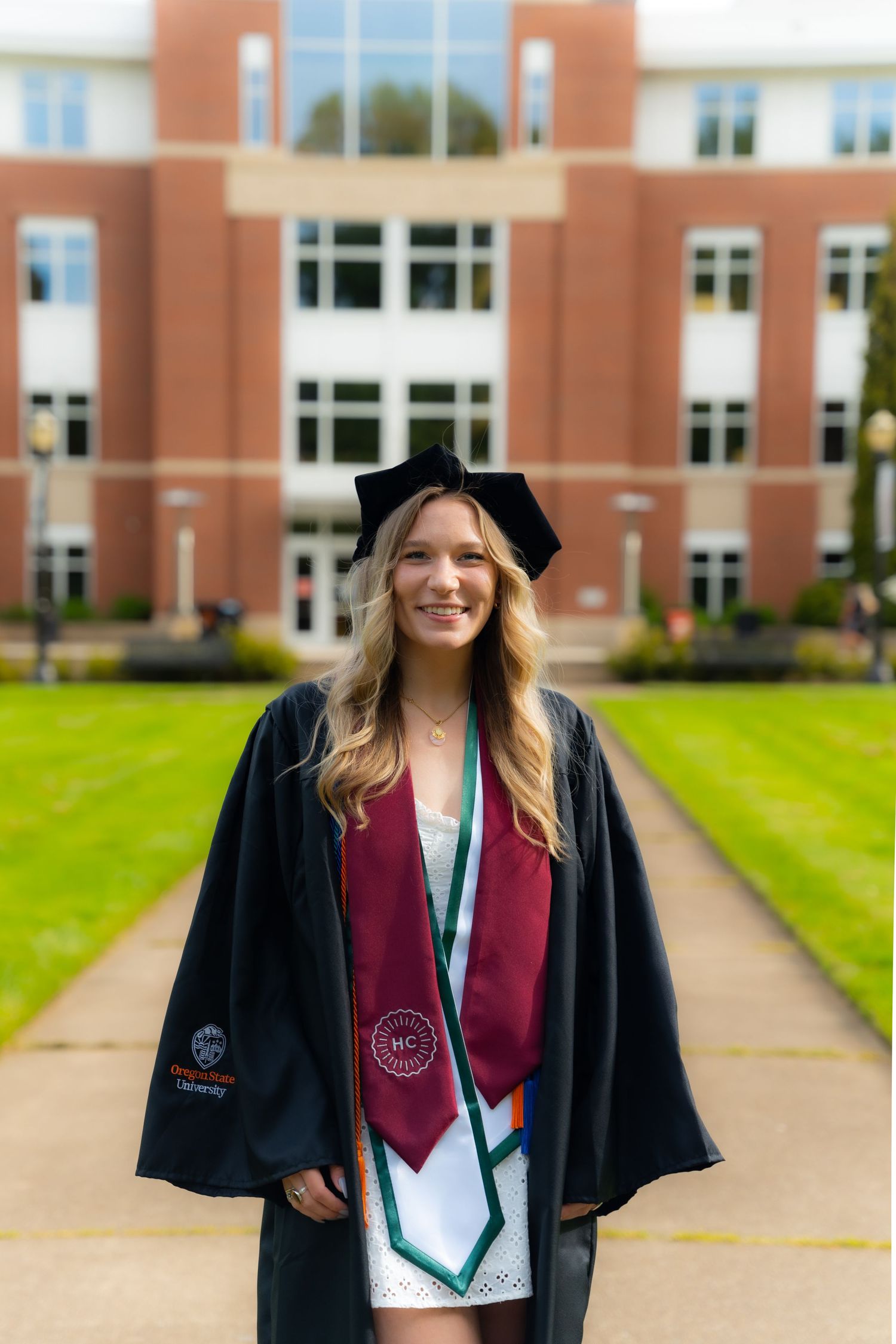 Graduate student in black cap and gown with burgundy stole stands smiling on campus walkway near university building.