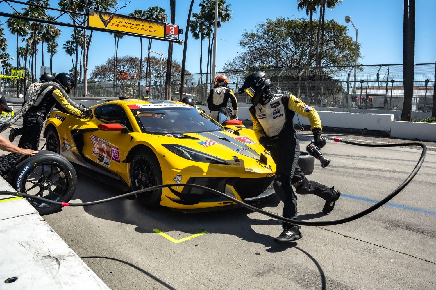 Yellow race car receiving a pit stop service with crew members during a motorsport competition on a sunny day.