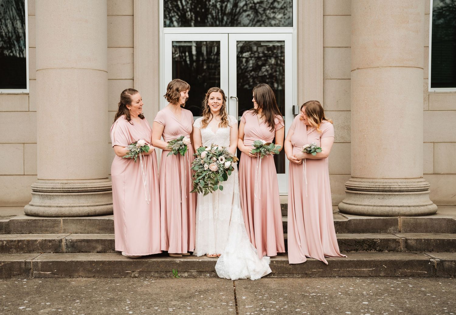 Bridal party in soft pink dresses stands near stone pillars at an elegant wedding venue.
