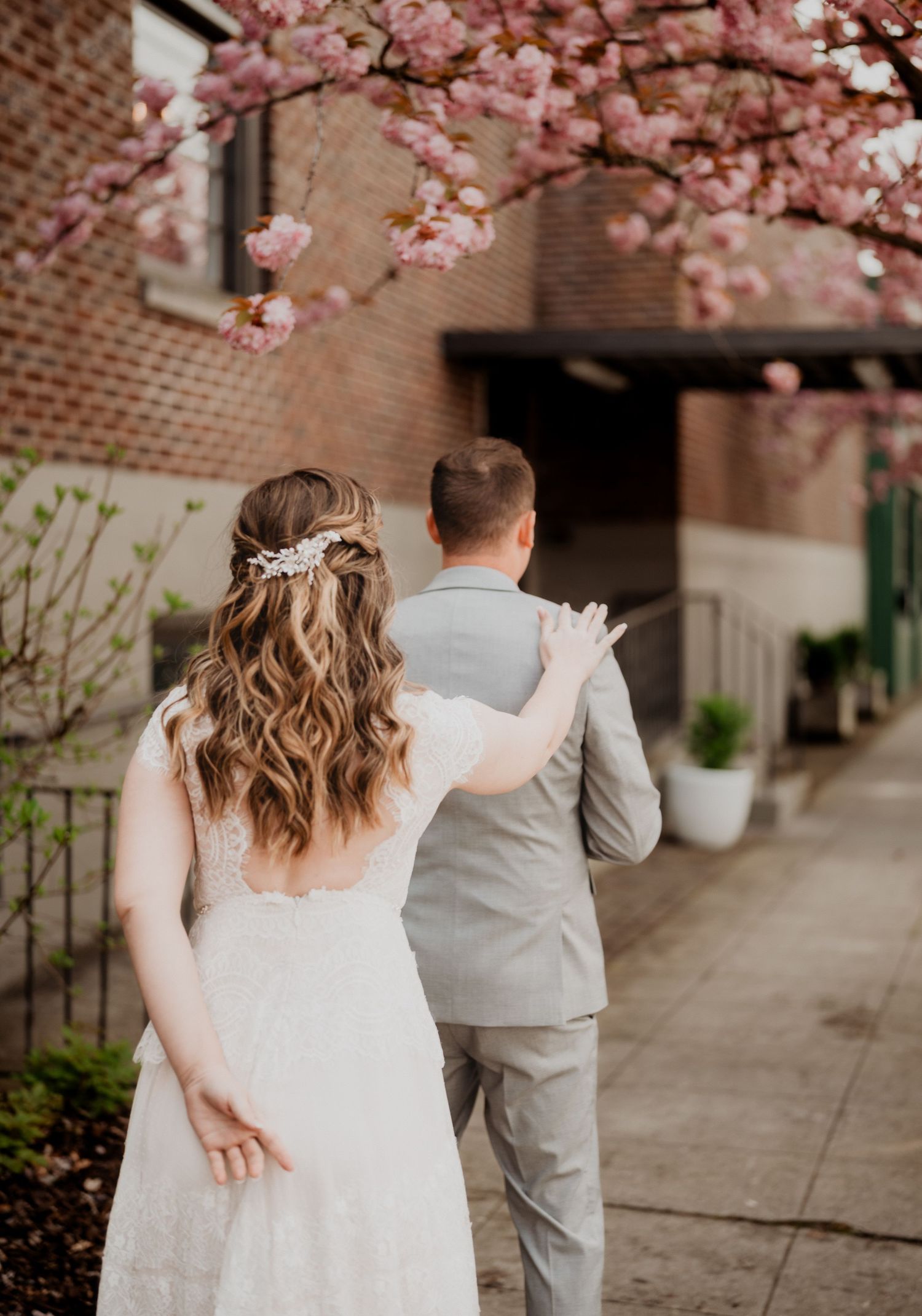 A romantic wedding moment under blooming cherry blossoms with bride in lace dress and groom in light gray suit.