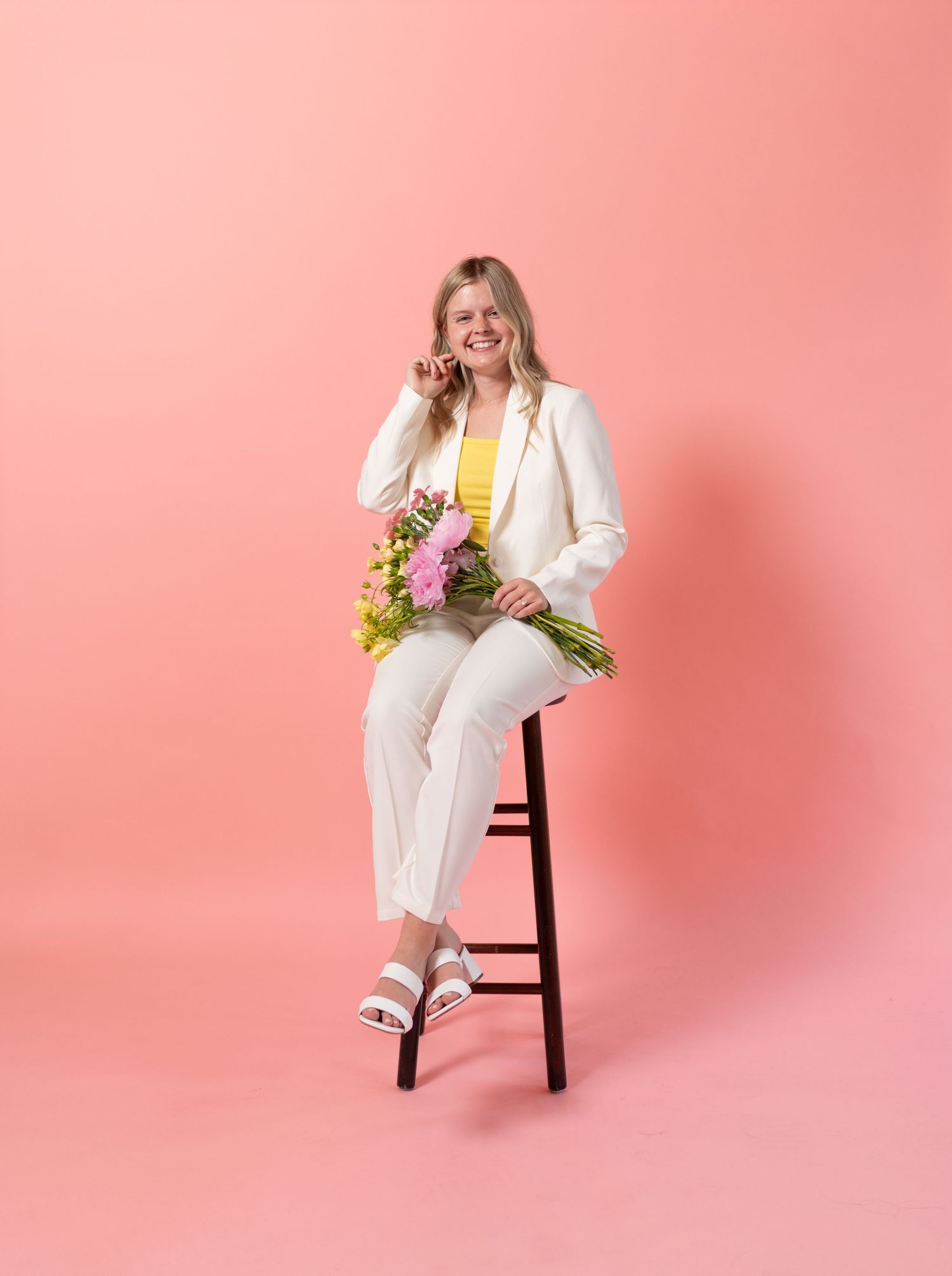 Person in white suit sitting on wooden stool against pink background holding colorful flowers.