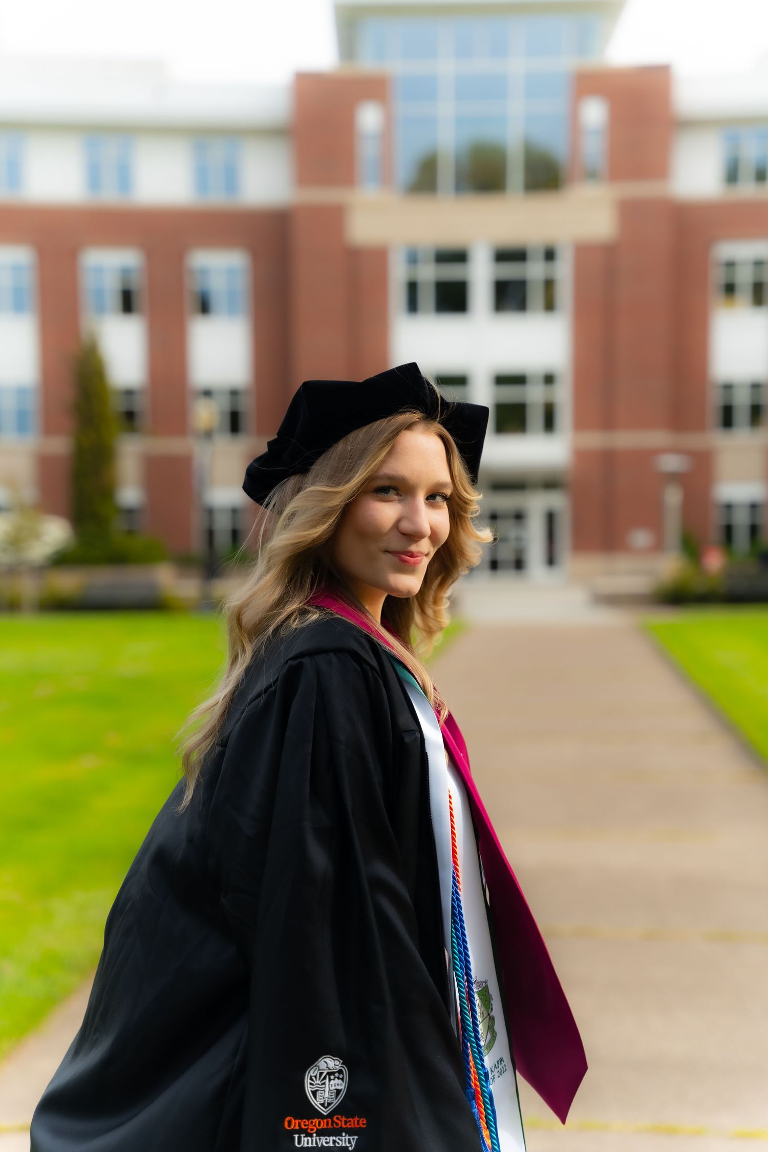 A graduate stands in front of a brick building on campus wearing a black cap and gown with a burgundy top beneath.