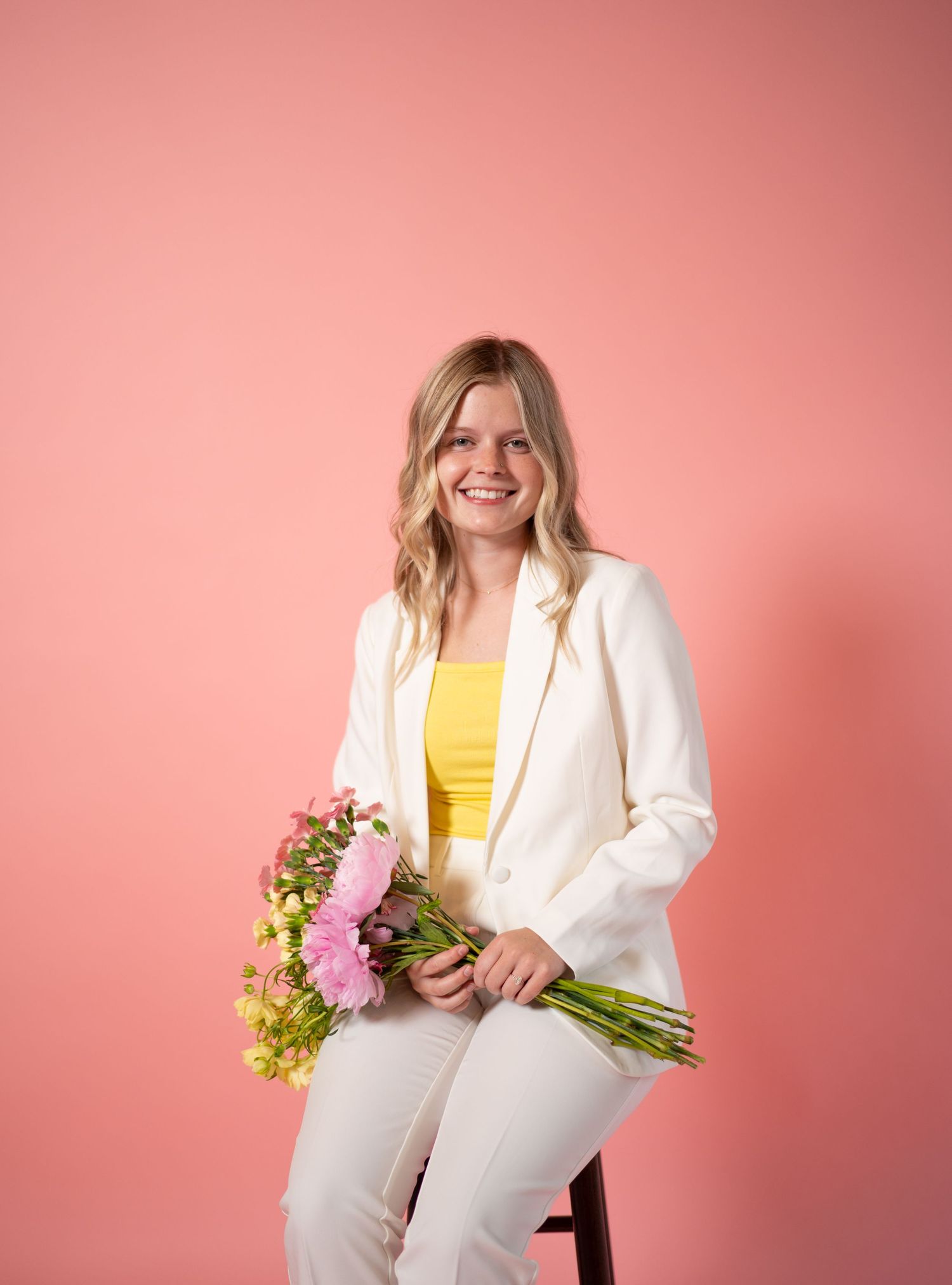 A person in a white suit and yellow top holds a bouquet of pink flowers against a pink background.