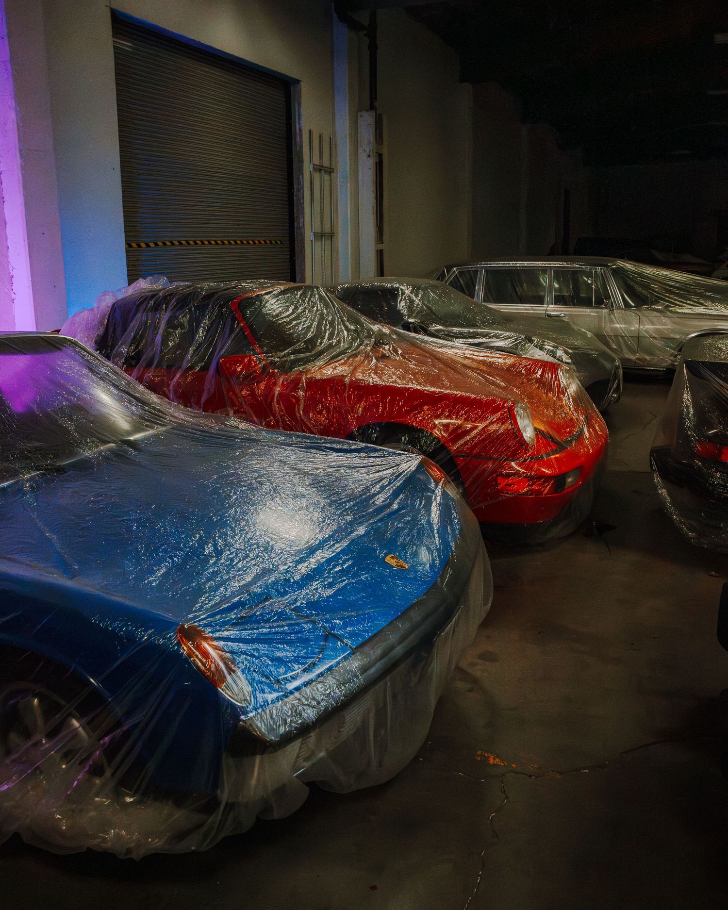 Luxury cars covered in protective plastic wraps are stored in a dimly lit garage with purple ambient lighting.