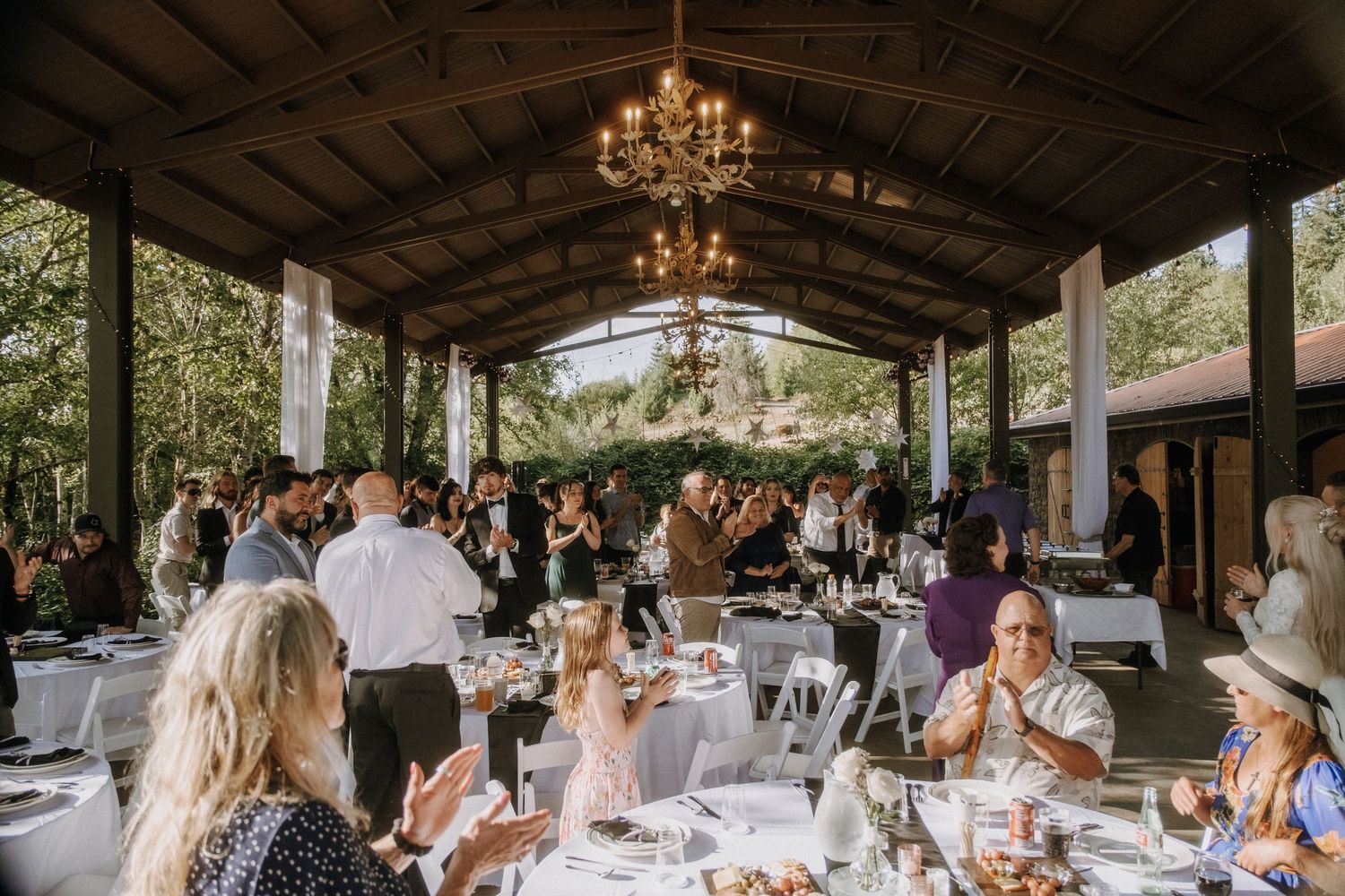 Wedding reception set under rustic wooden pavilion with chandelier lighting.