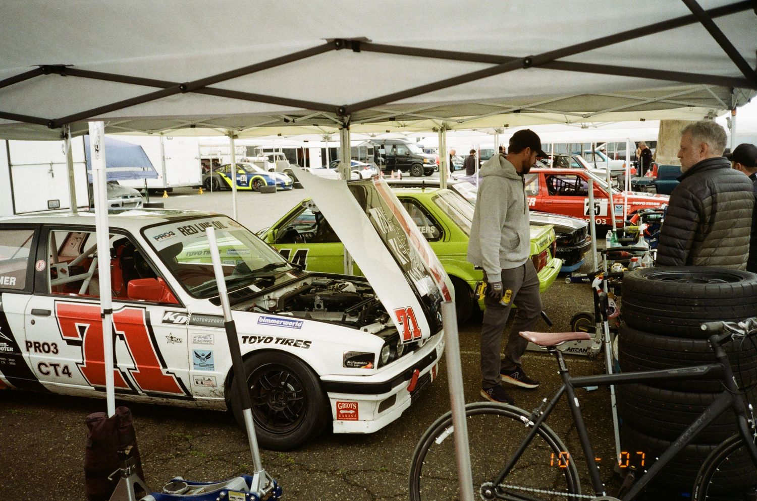 Classic rally car with number 57 and red stripes parked under tent canopy at motorsport event with spectators.