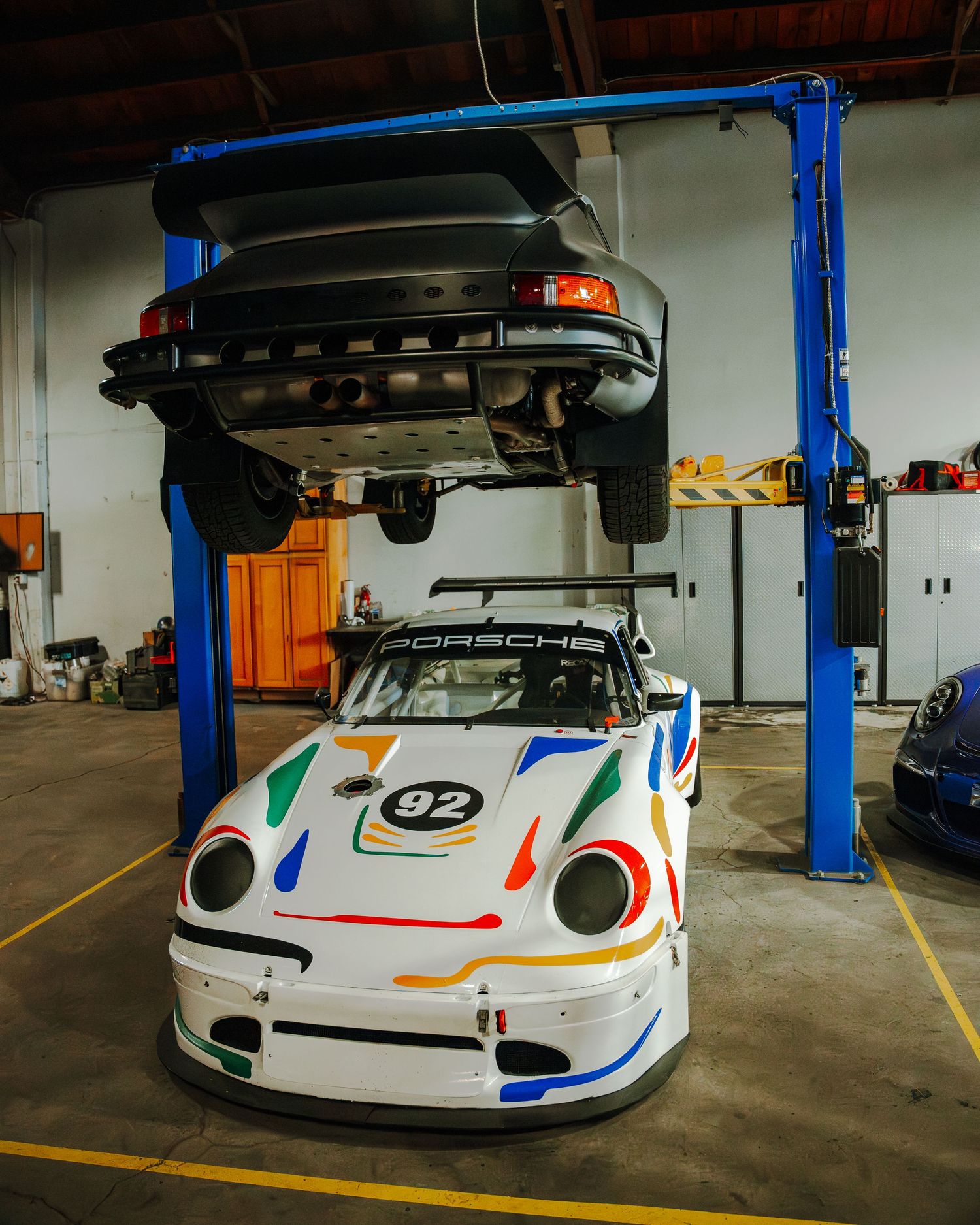 White Porsche 911 GT3R race car being serviced on a hydraulic lift in an auto workshop garage.