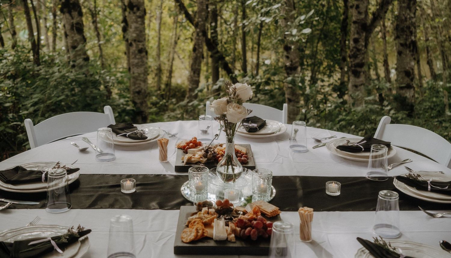 An elegant outdoor dining table set in a forest with wine glasses, charcuterie boards, and natural decor.