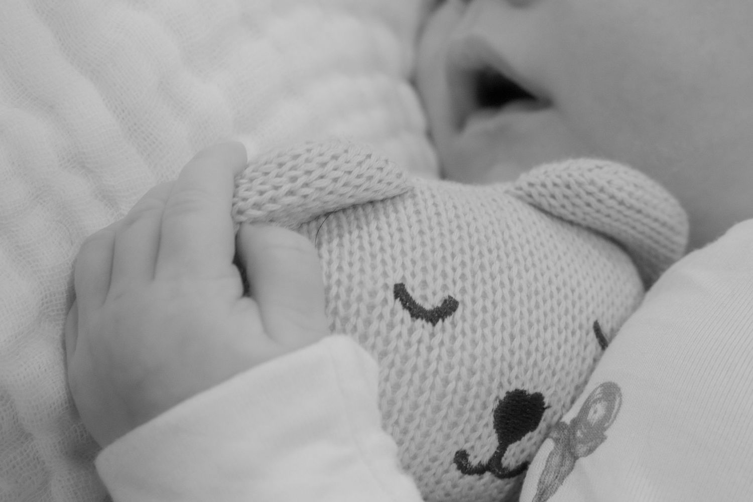 Black and white close-up of knitted plush toy held by newborn on bedding.