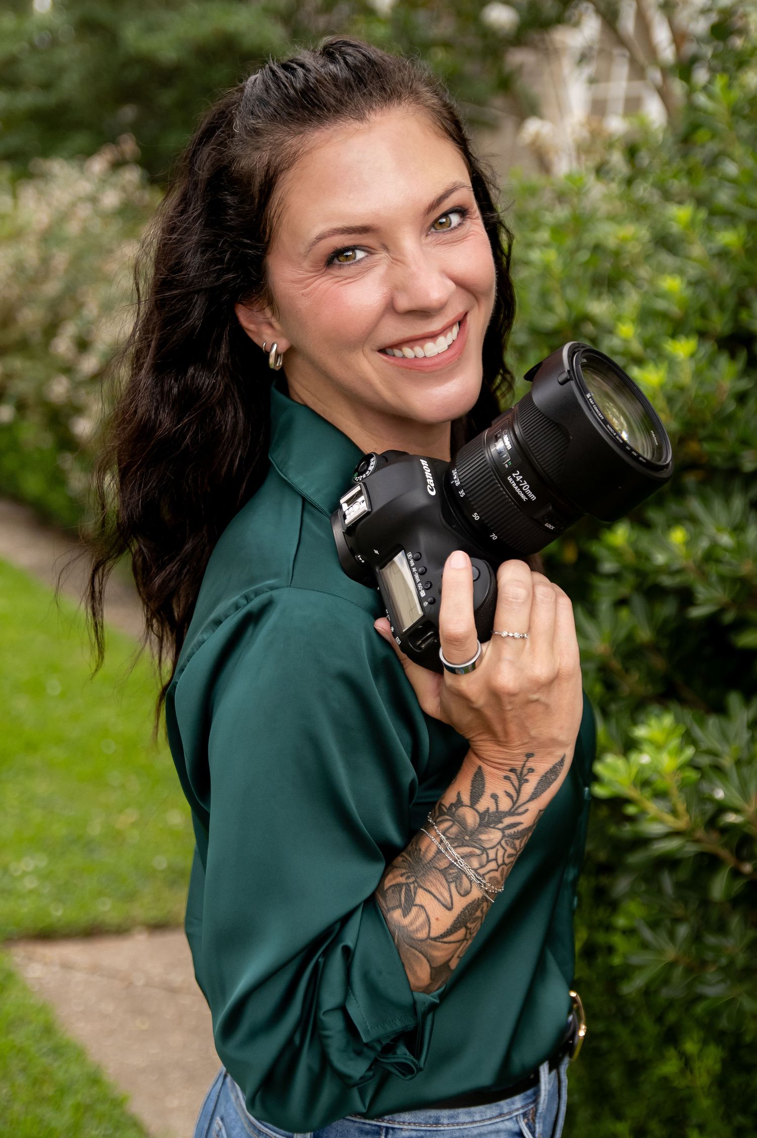 Professional photographer in emerald blouse poses with camera in outdoor garden setting.