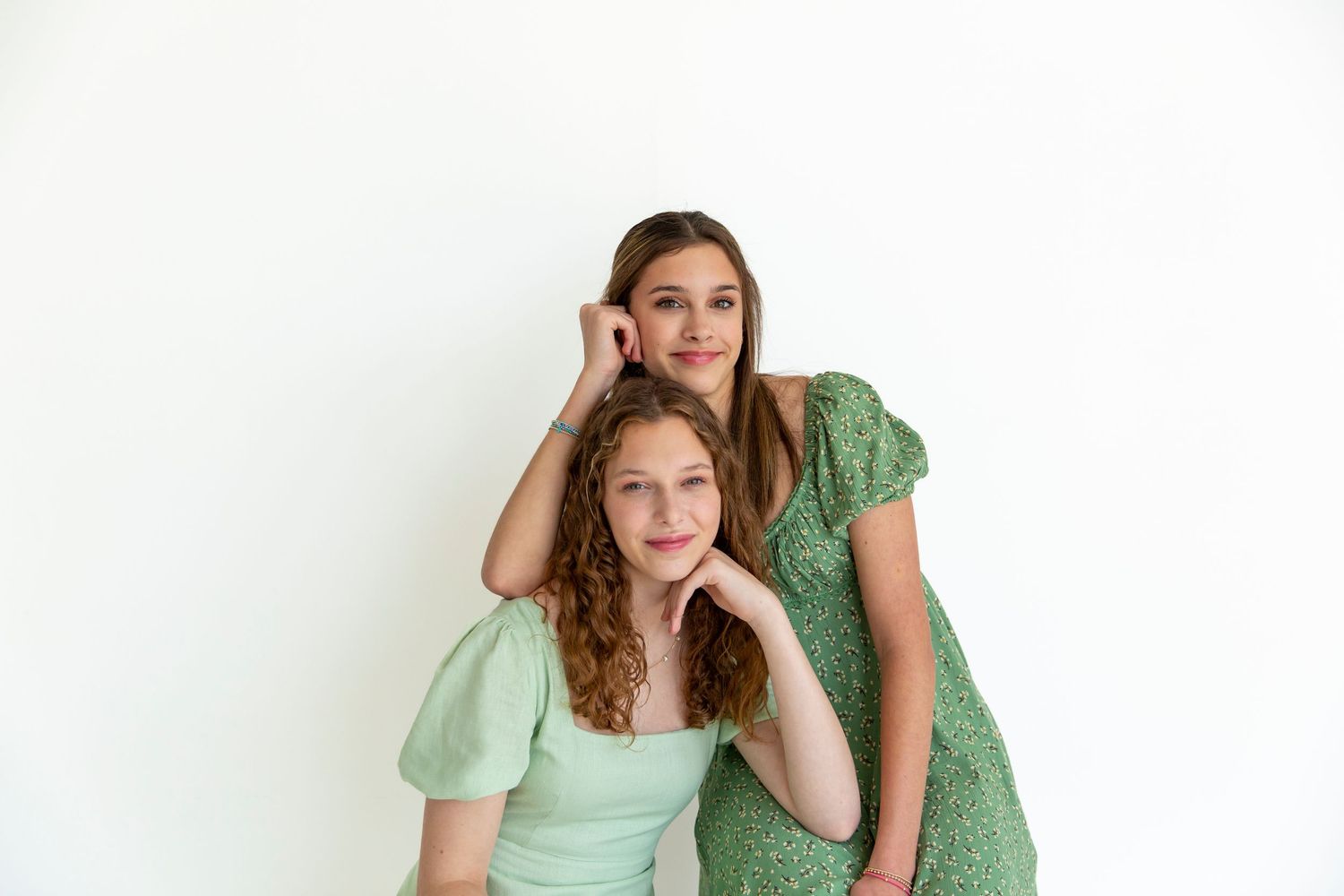 Two women in green dresses lean close together for a portrait against a white wall.