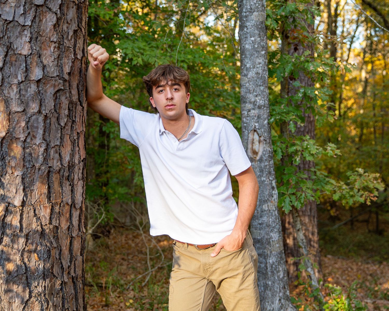 A person in a white shirt and khaki pants poses casually against a tree trunk in a wooded outdoor setting.