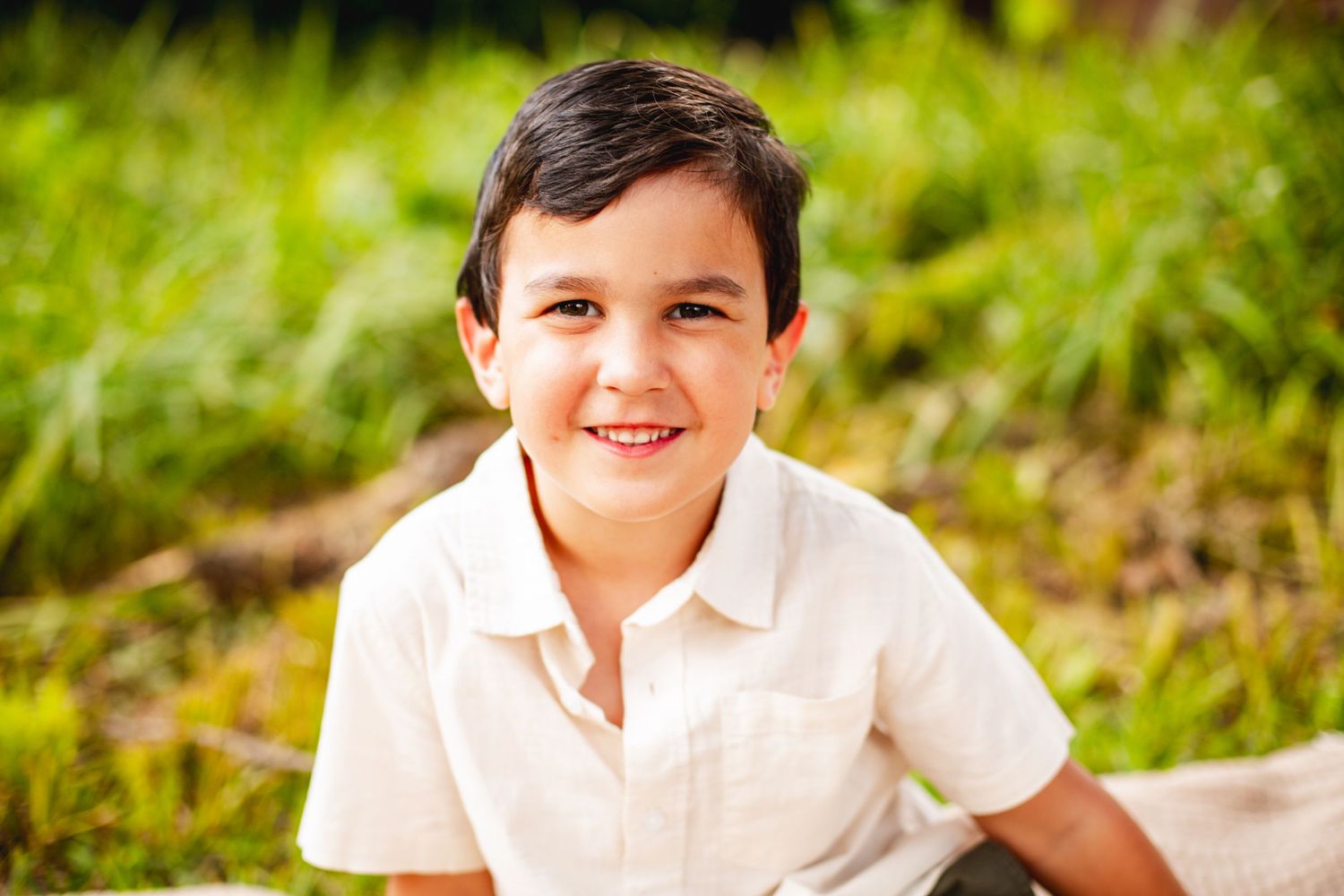 Child in white polo shirt sitting outdoors on grass in natural lighting.