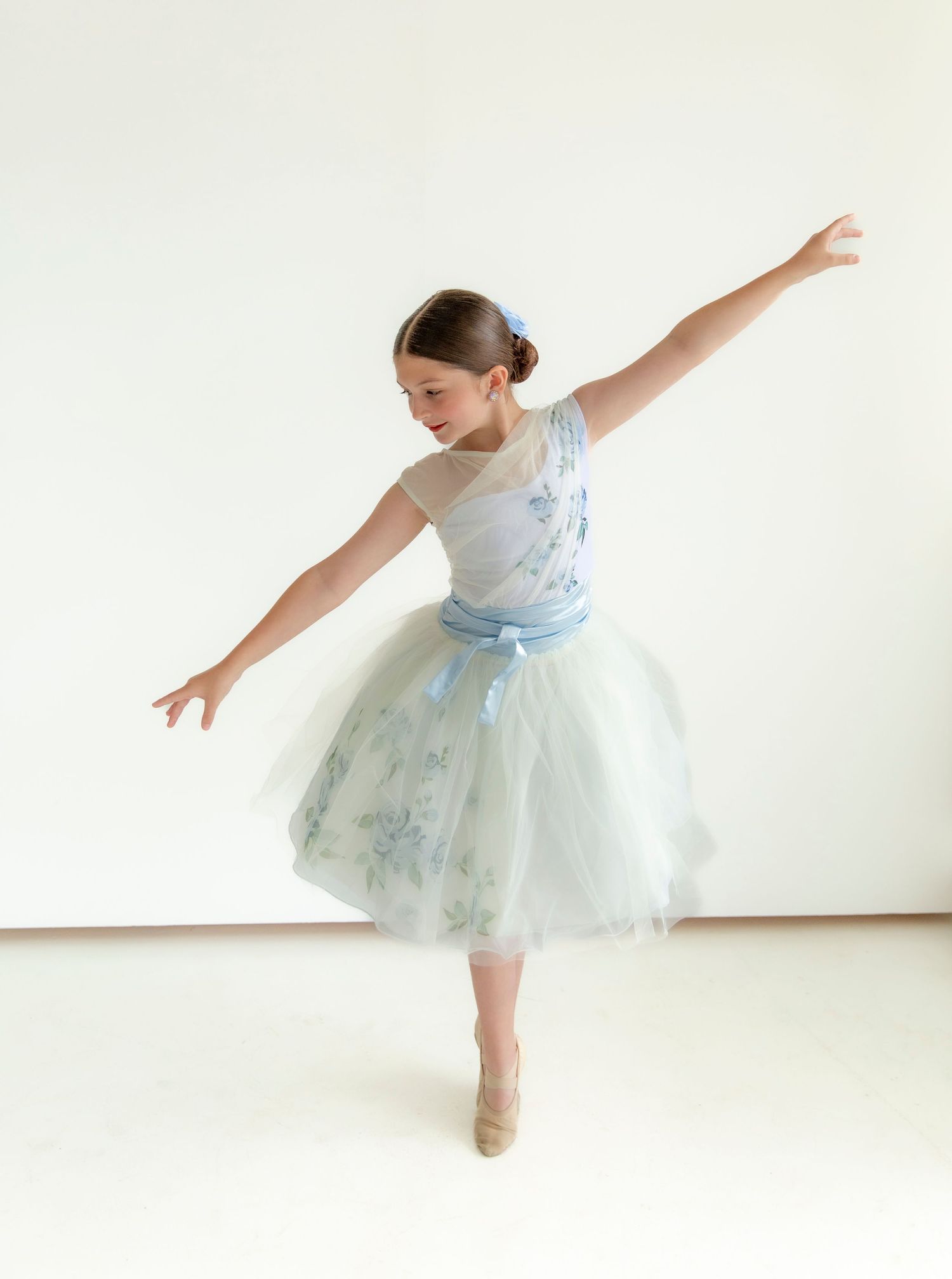 Young dancer in light blue tulle dress performs graceful movements against white background.