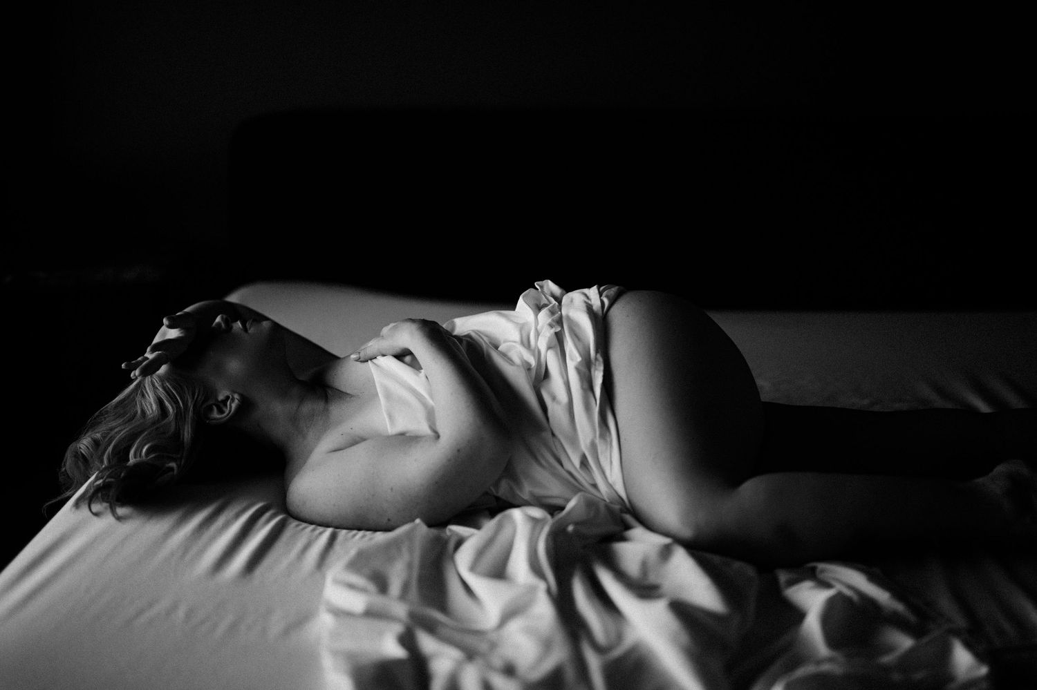 A dramatic black and white artistic photograph of a person reclining on light colored bedding with strong shadows.