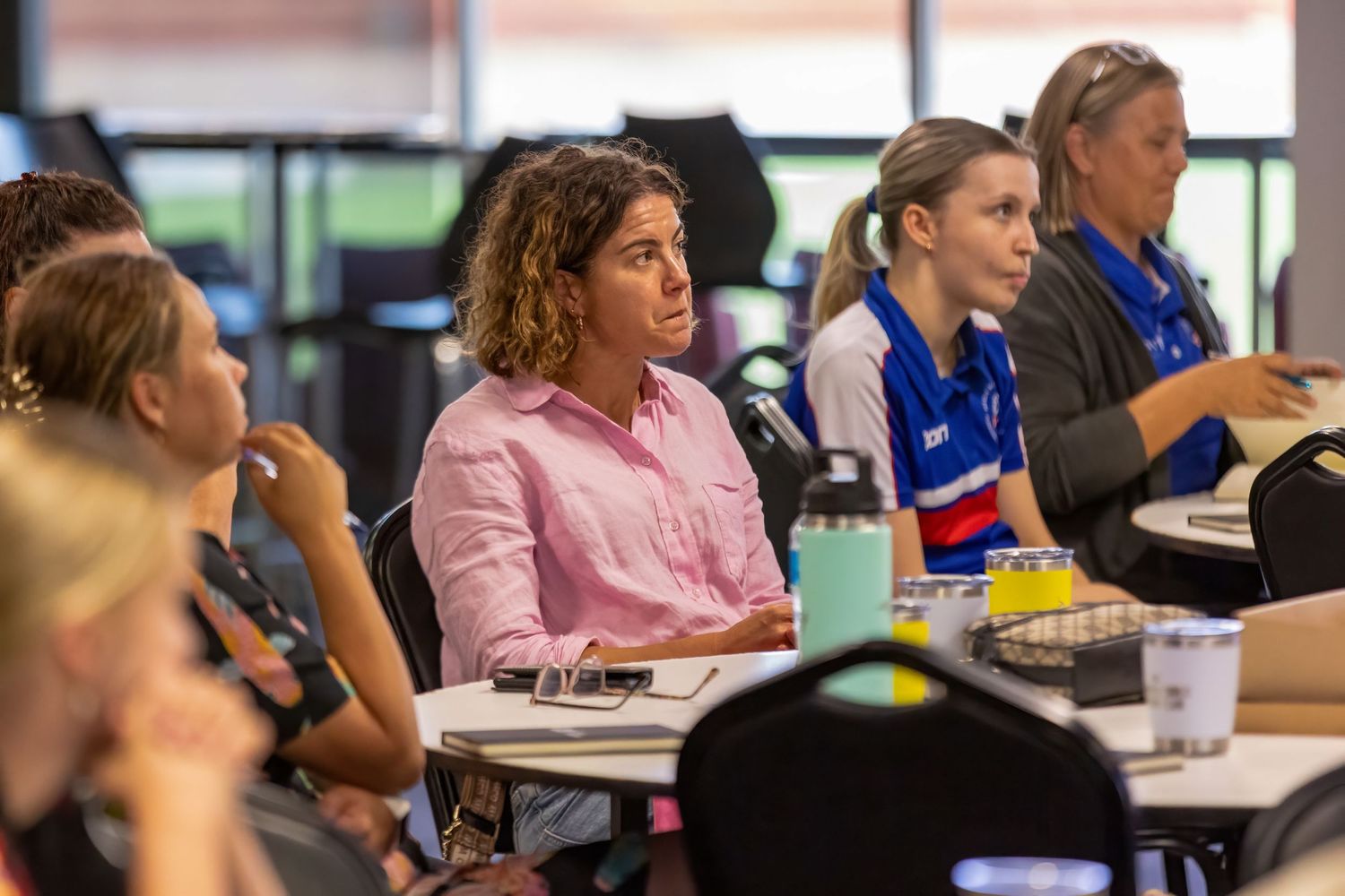 Students in a classroom setting sit at desks during a group discussion or presentation.