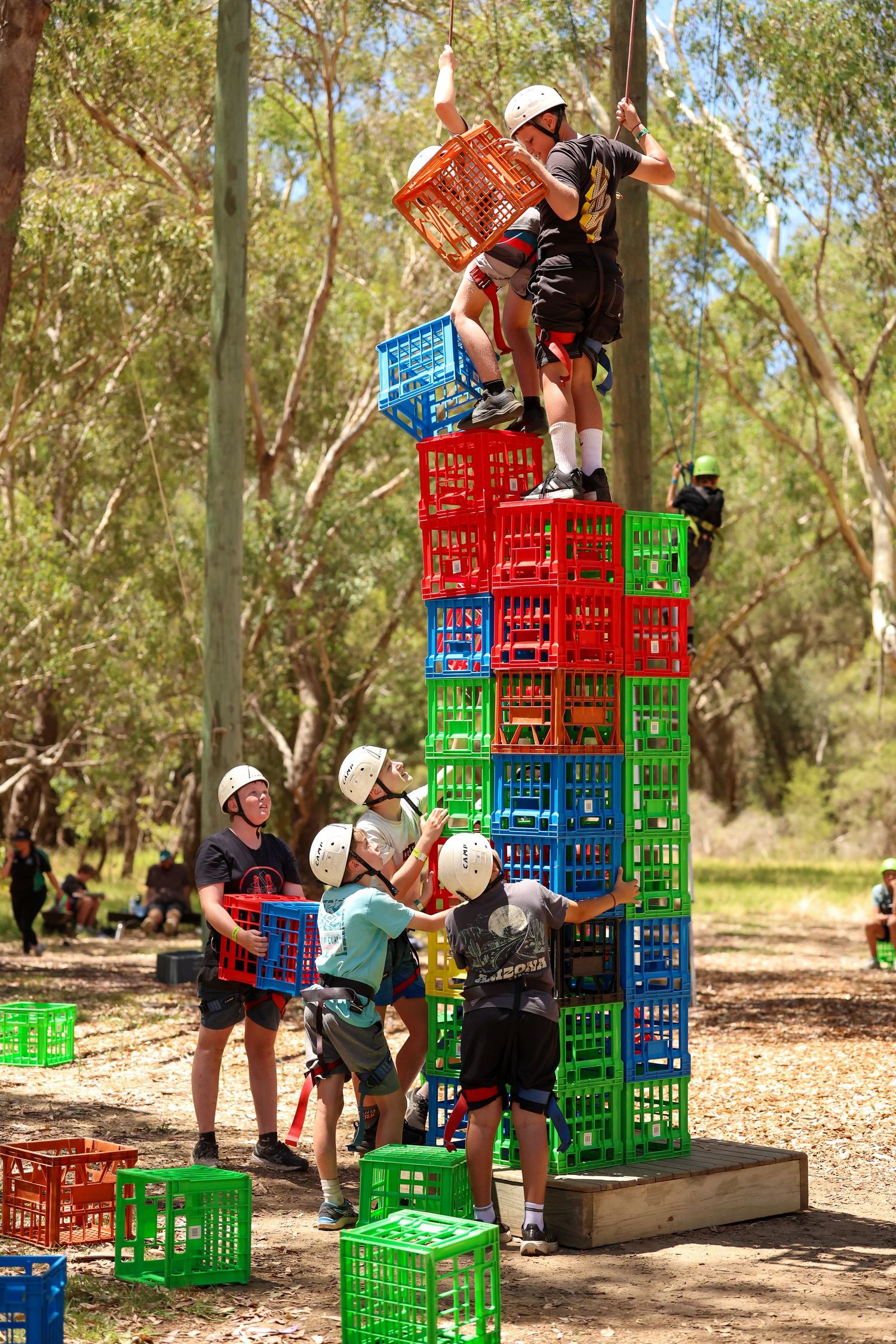 People stacking colorful milk crates outdoors in a team building challenge activity at a wooded park area.