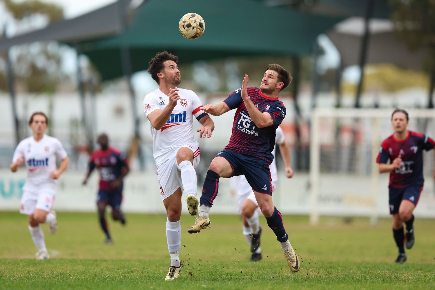 Two soccer players jump high to compete for the ball during an intense match on a grass field.