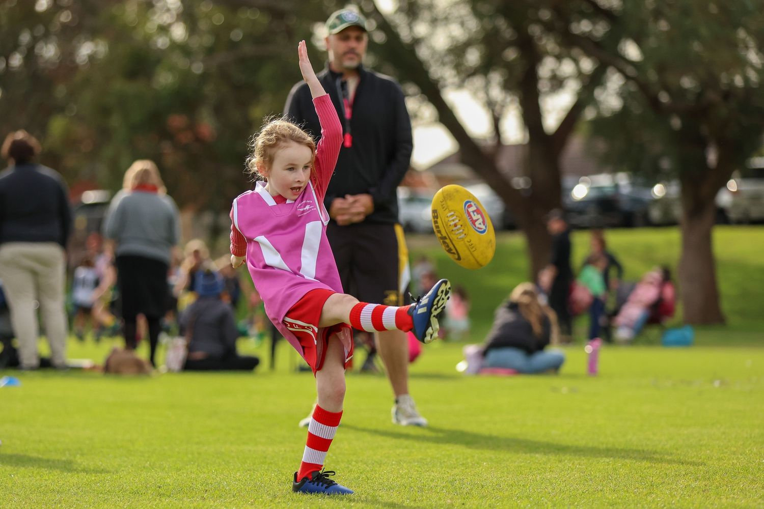 Young child in bright pink uniform kicking yellow football on grass field during community sports event.
