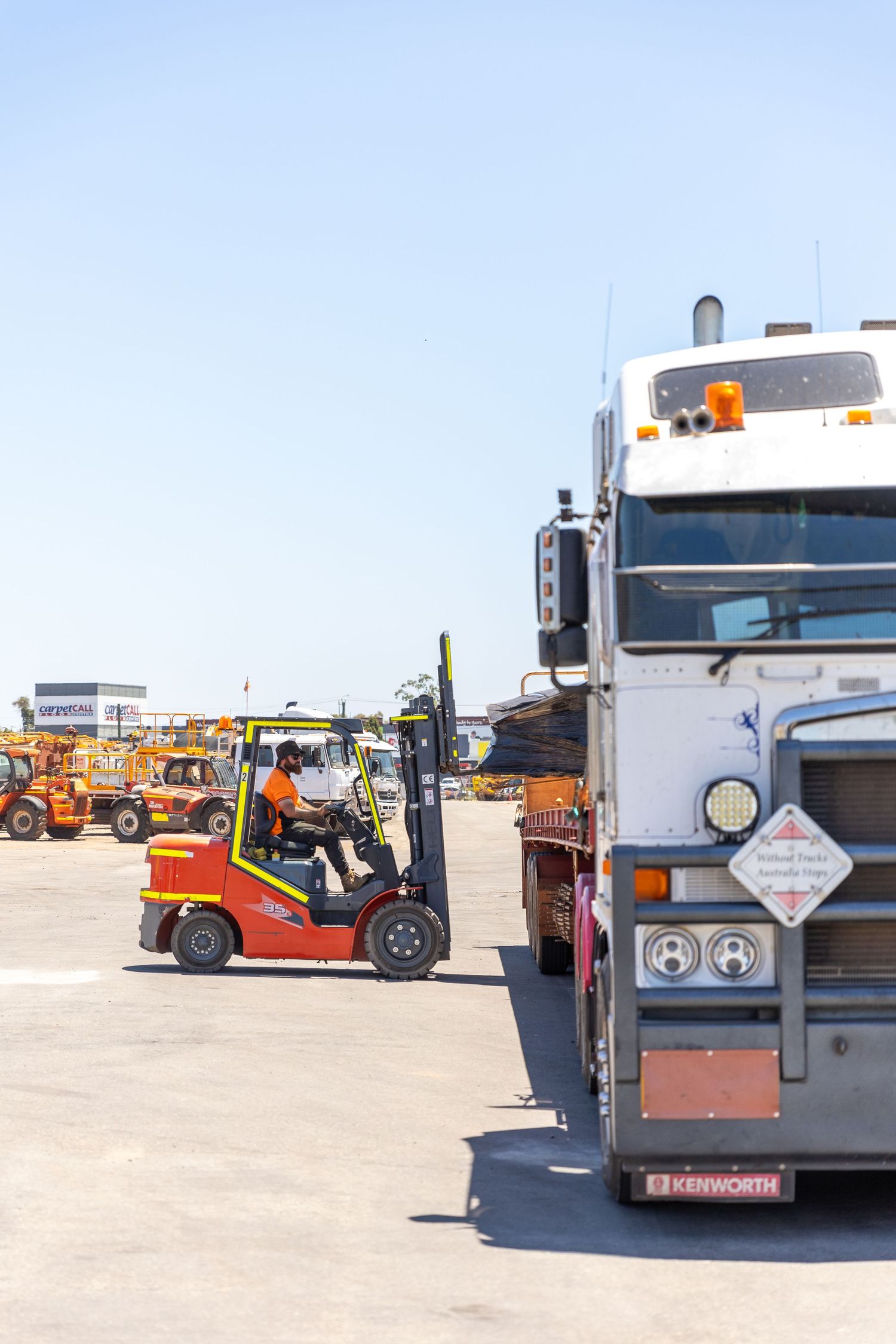 Heavy duty trucks and forklifts are parked in an industrial yard with clear blue sky in the background.