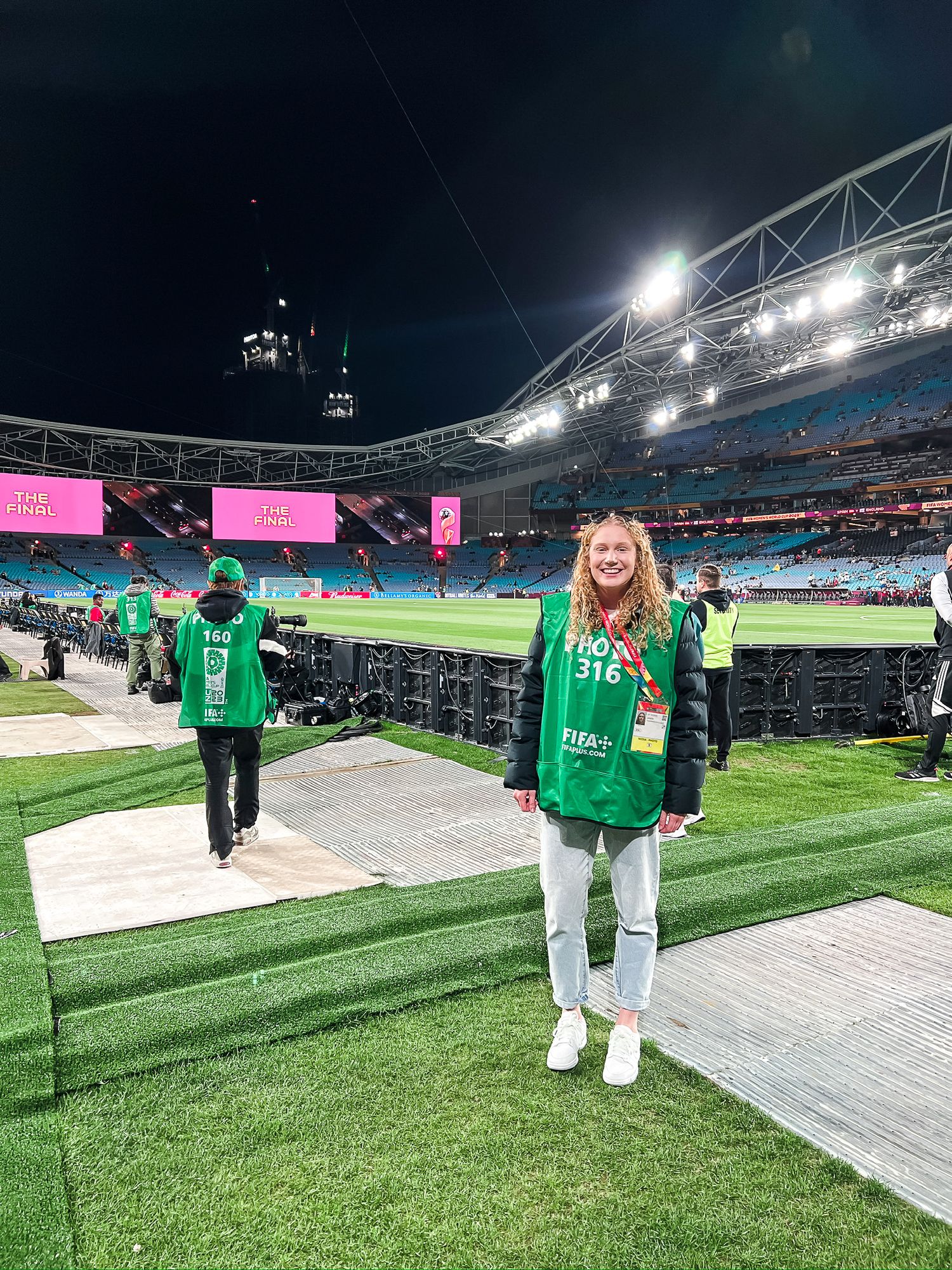 Event staff wearing green vests work on field at illuminated sports stadium at night.
