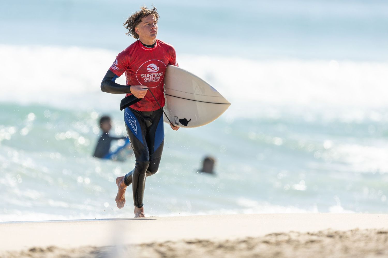 A surfer in a red t-shirt carries a white surfboard across a sandy beach with ocean waves in the background.