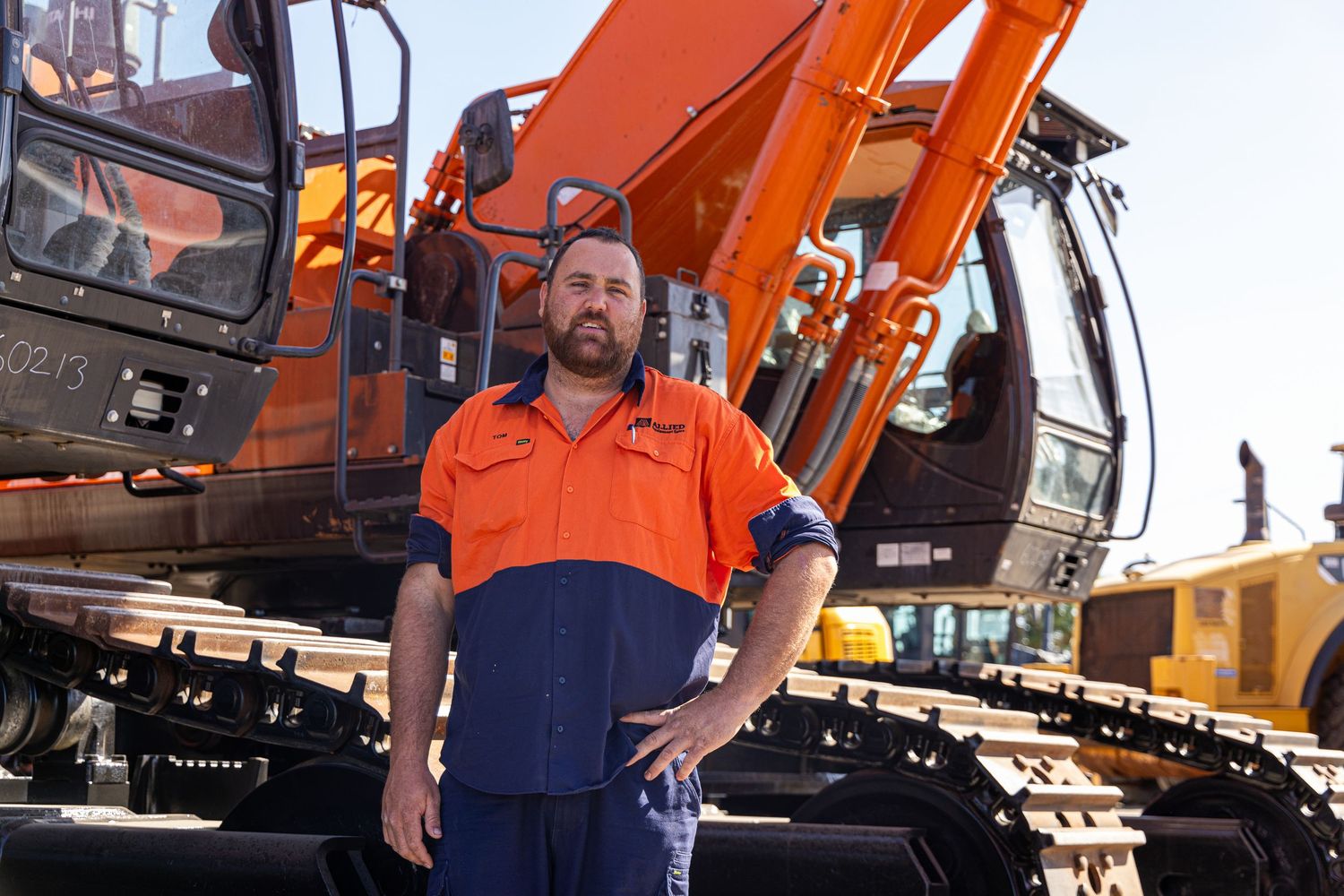 Construction worker in orange and navy hi-vis uniform standing in front of large orange excavator machinery.