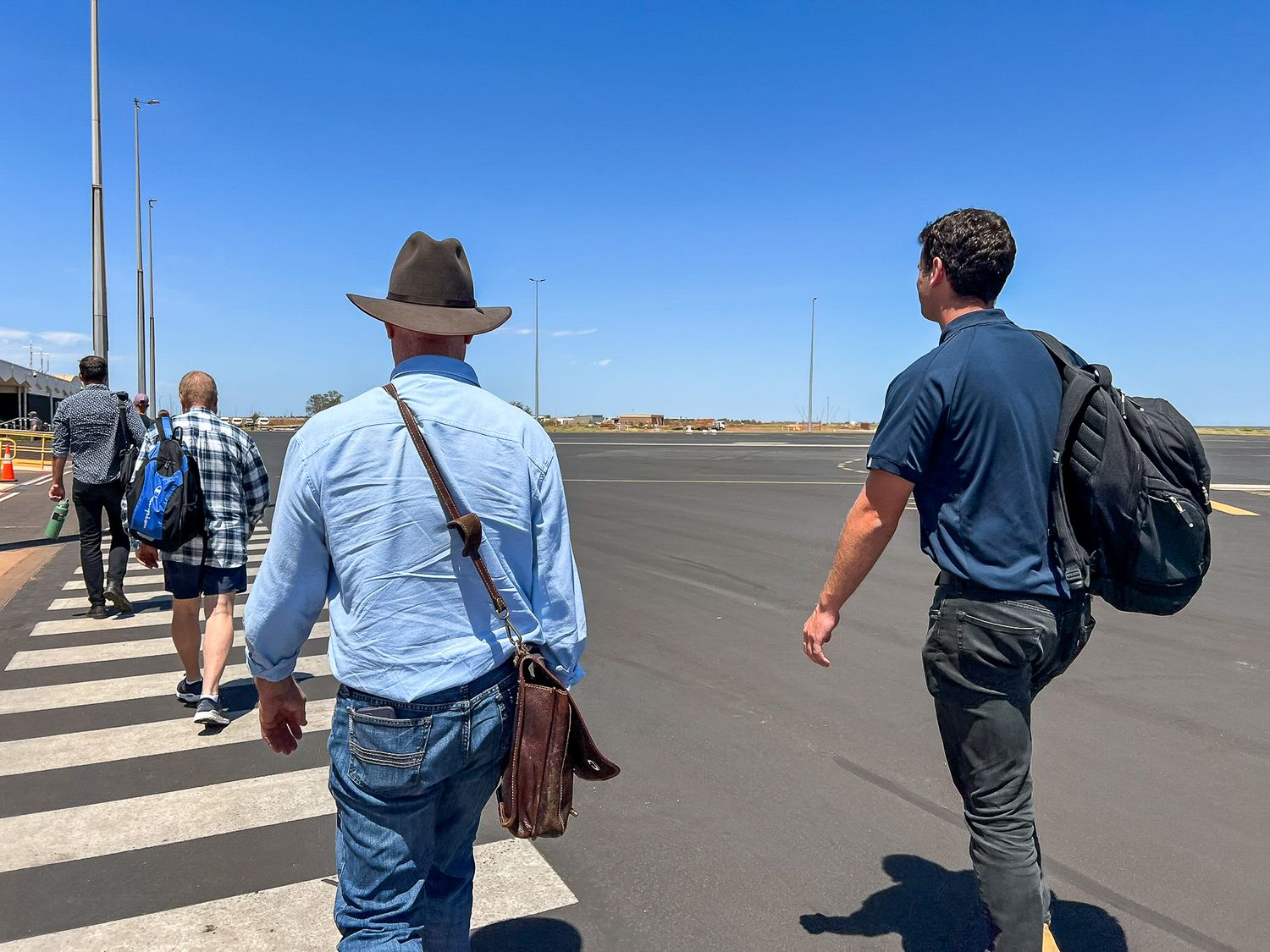 Passengers walking across an airport tarmac carrying backpacks and bags under a bright blue sky.