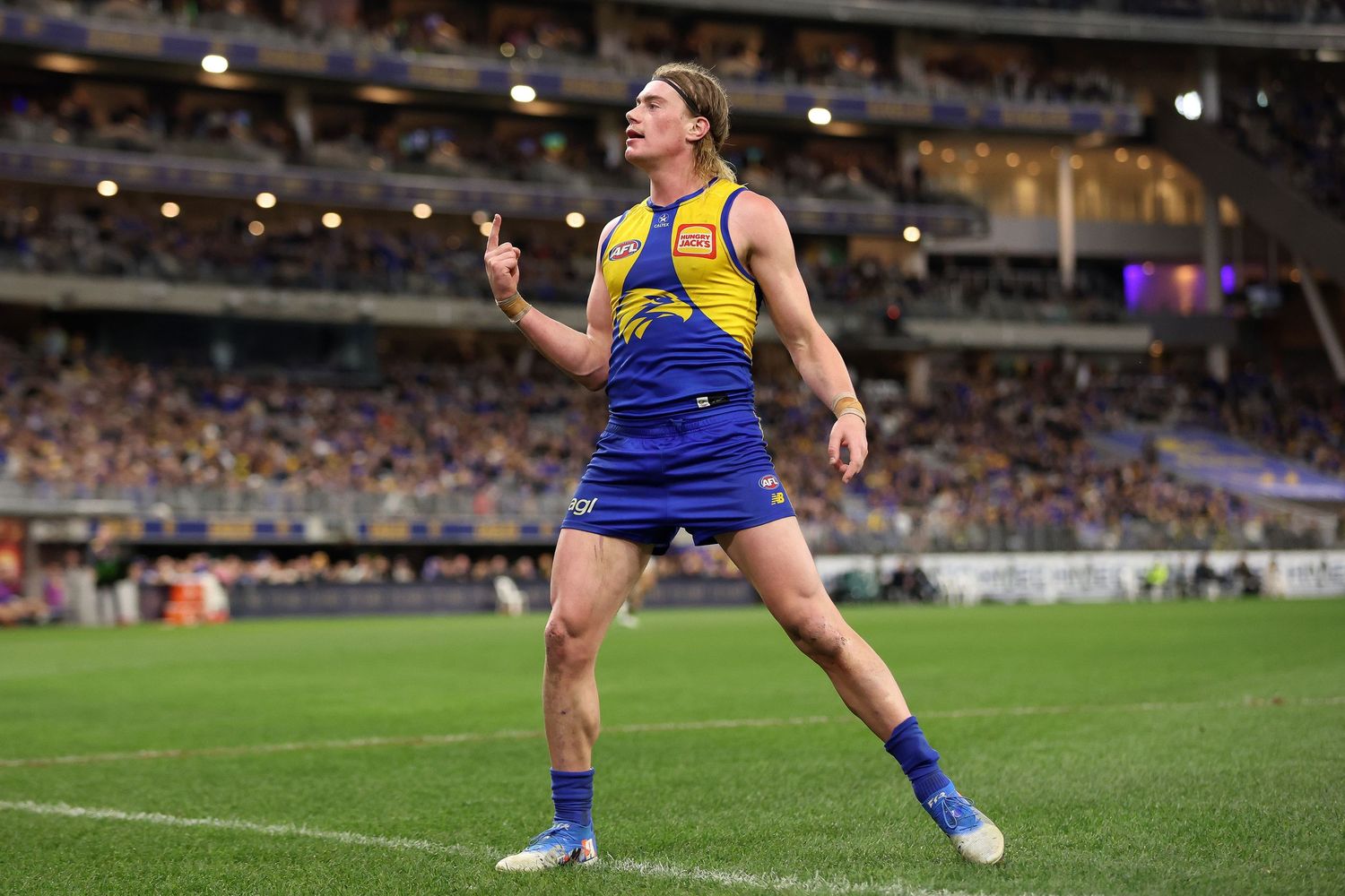 AFL football player in West Coast Eagles uniform celebrates on field at night stadium during match.
