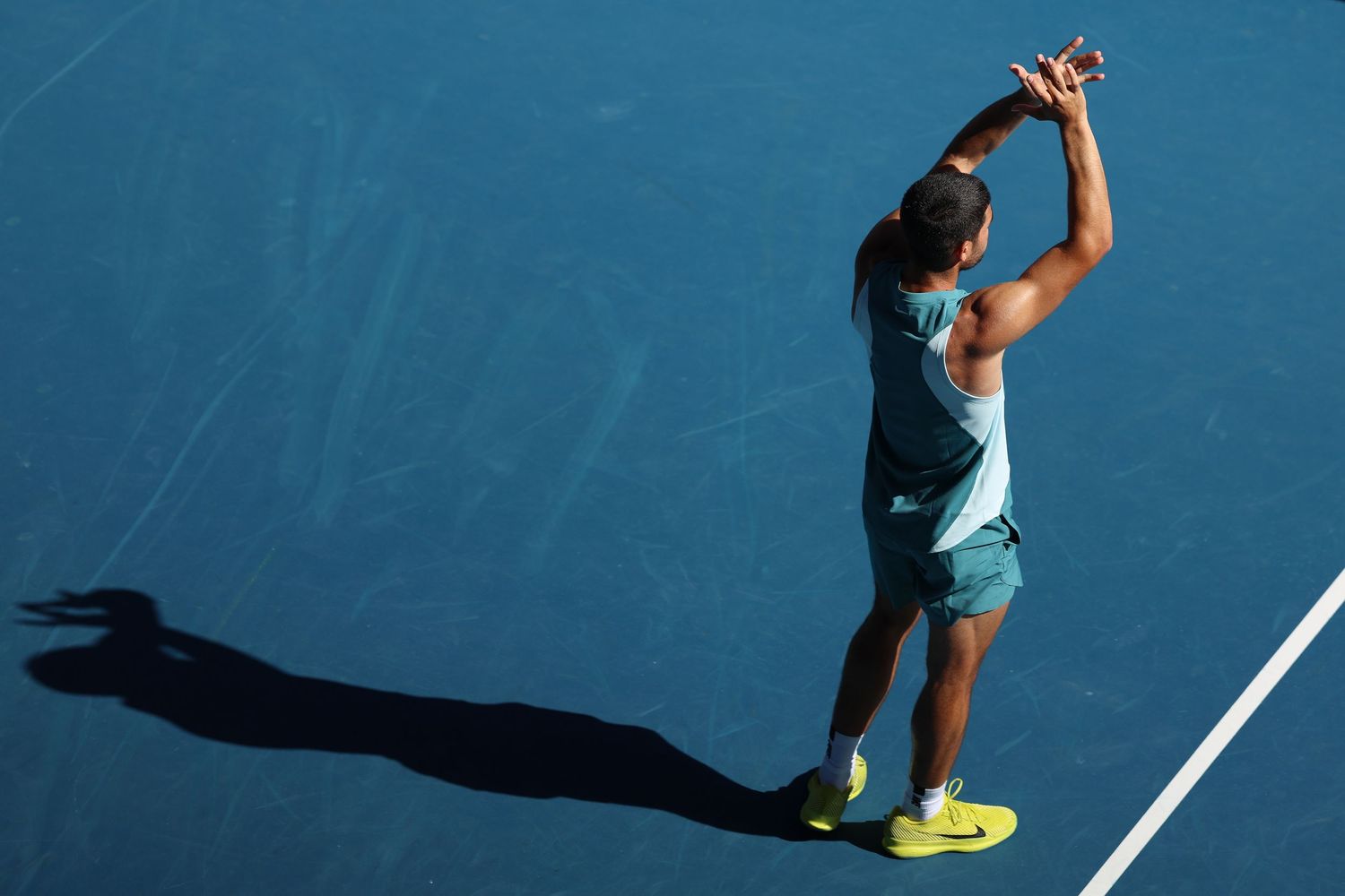 Tennis player serves ball on sunny blue court creating dramatic shadow.
