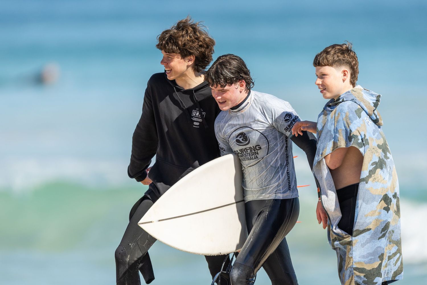 A group of surfers carrying surfboards walk along the beach in casual attire with a blue ocean backdrop.