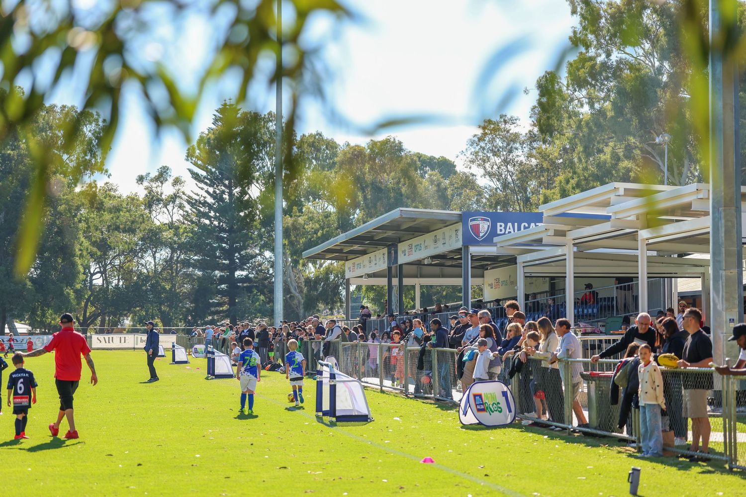Crowd of spectators lining the fence at Balcatta oval watching an outdoor Australian rules football event on a sunny day.