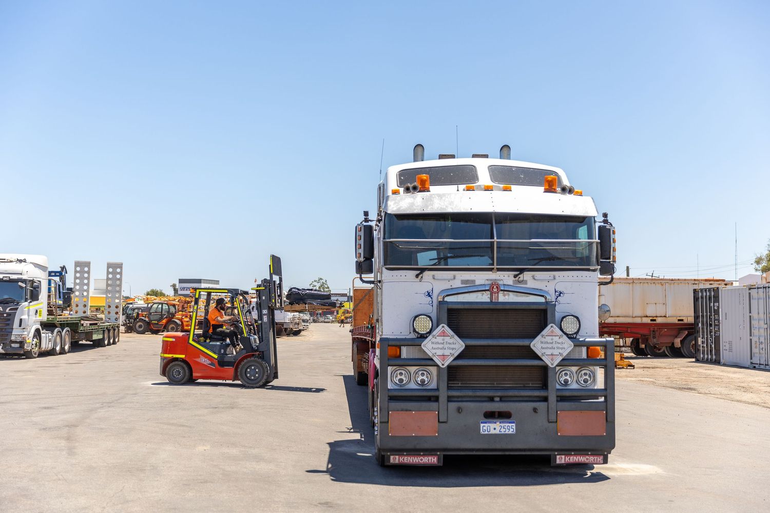 Front view of a Kenworth semi truck with forklift in background at an industrial yard on a sunny day.