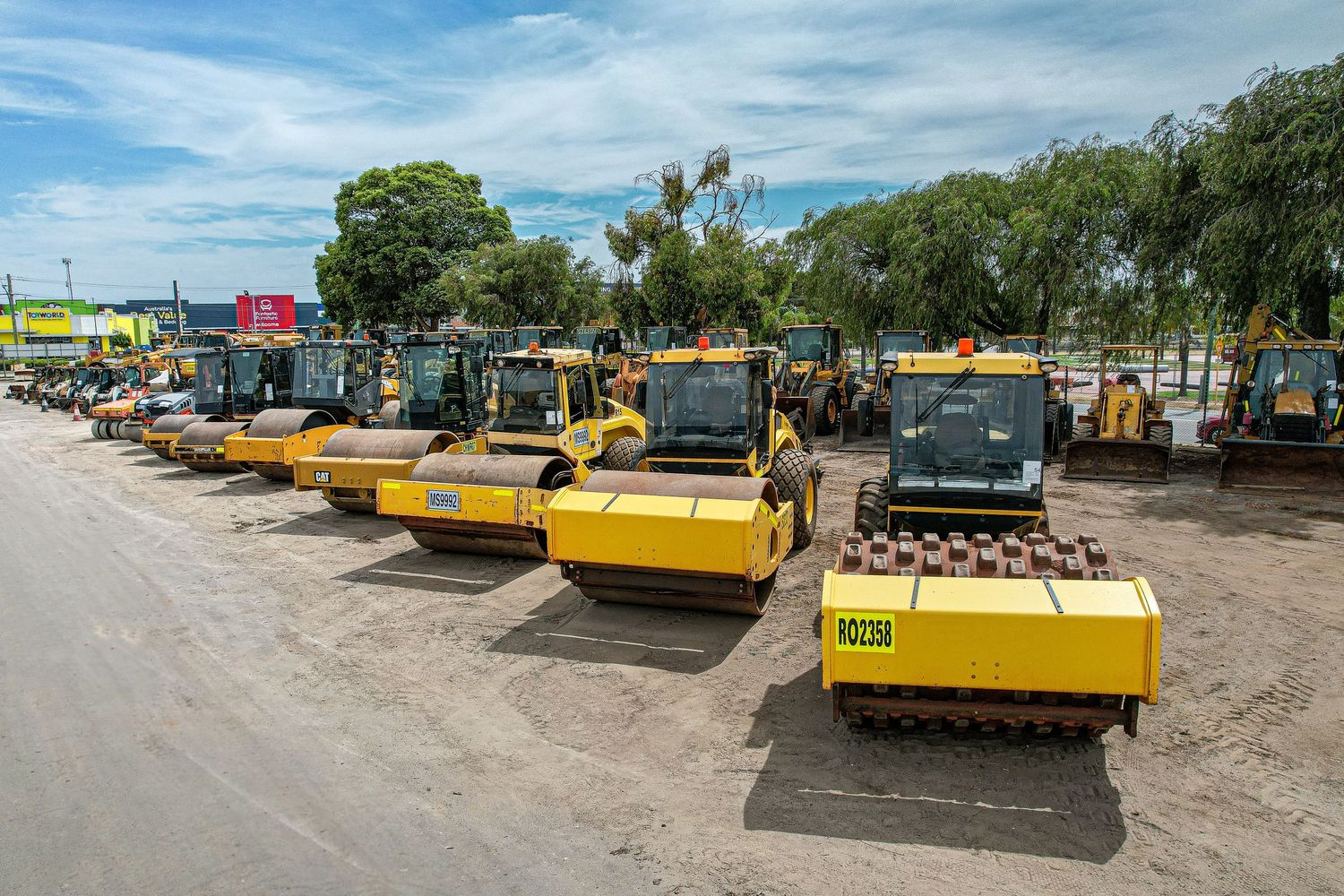 A row of yellow construction rollers and heavy machinery parked in a lot under blue skies with trees in background.