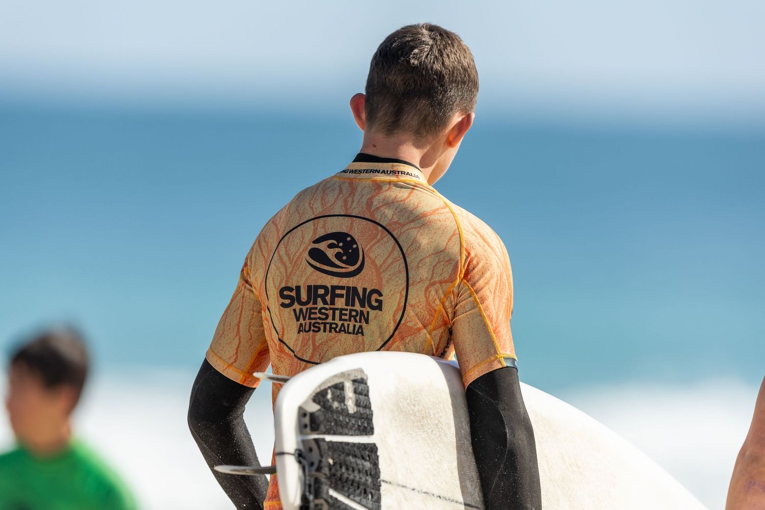 Surfers walking along a beach break carrying surfboards and wearing wetsuits on a sunny day with blue ocean waves.