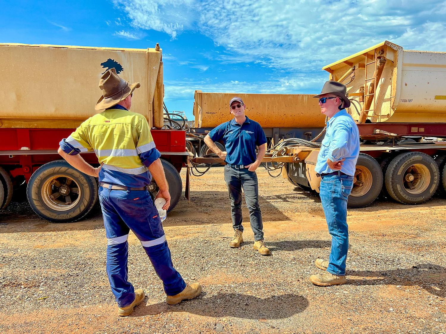 Group of workers having a discussion near yellow heavy machinery and transport trucks on a dusty worksite.